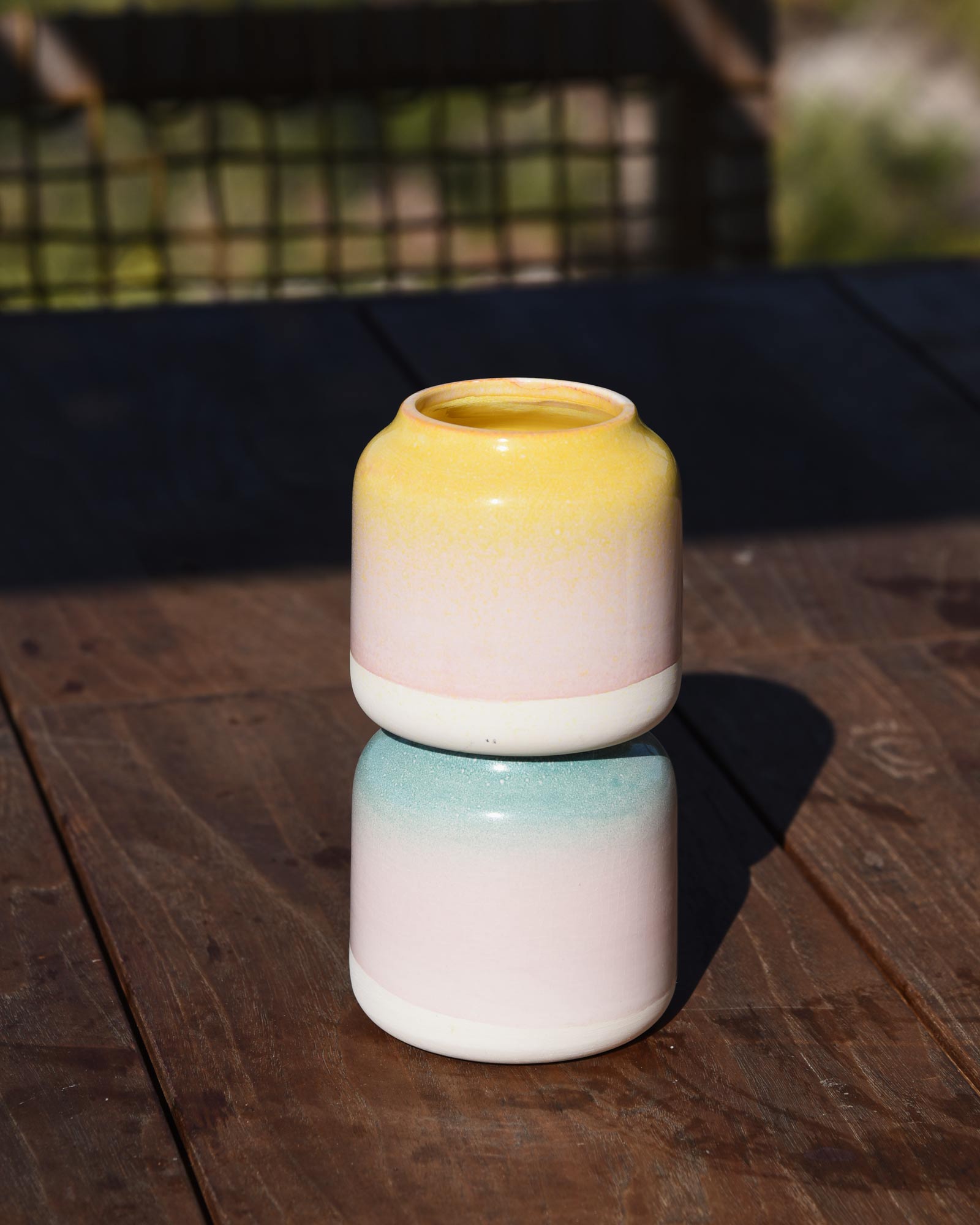 Zwei bunt glasierten Keramiktöpfen in Gelb, Rosa und Blau stehen gestapelt auf einem Holztisch in der Sonne.