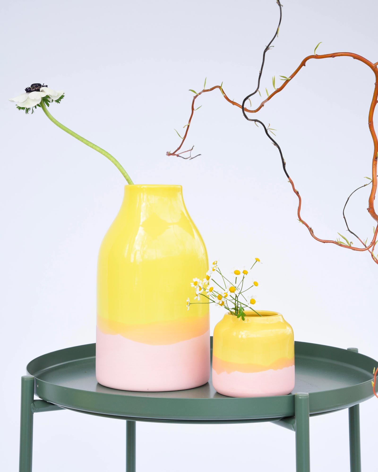 Zwei bunte Vasen mit Blumen auf einem runden, grünen Tisch vor einem hellen Hintergrund.