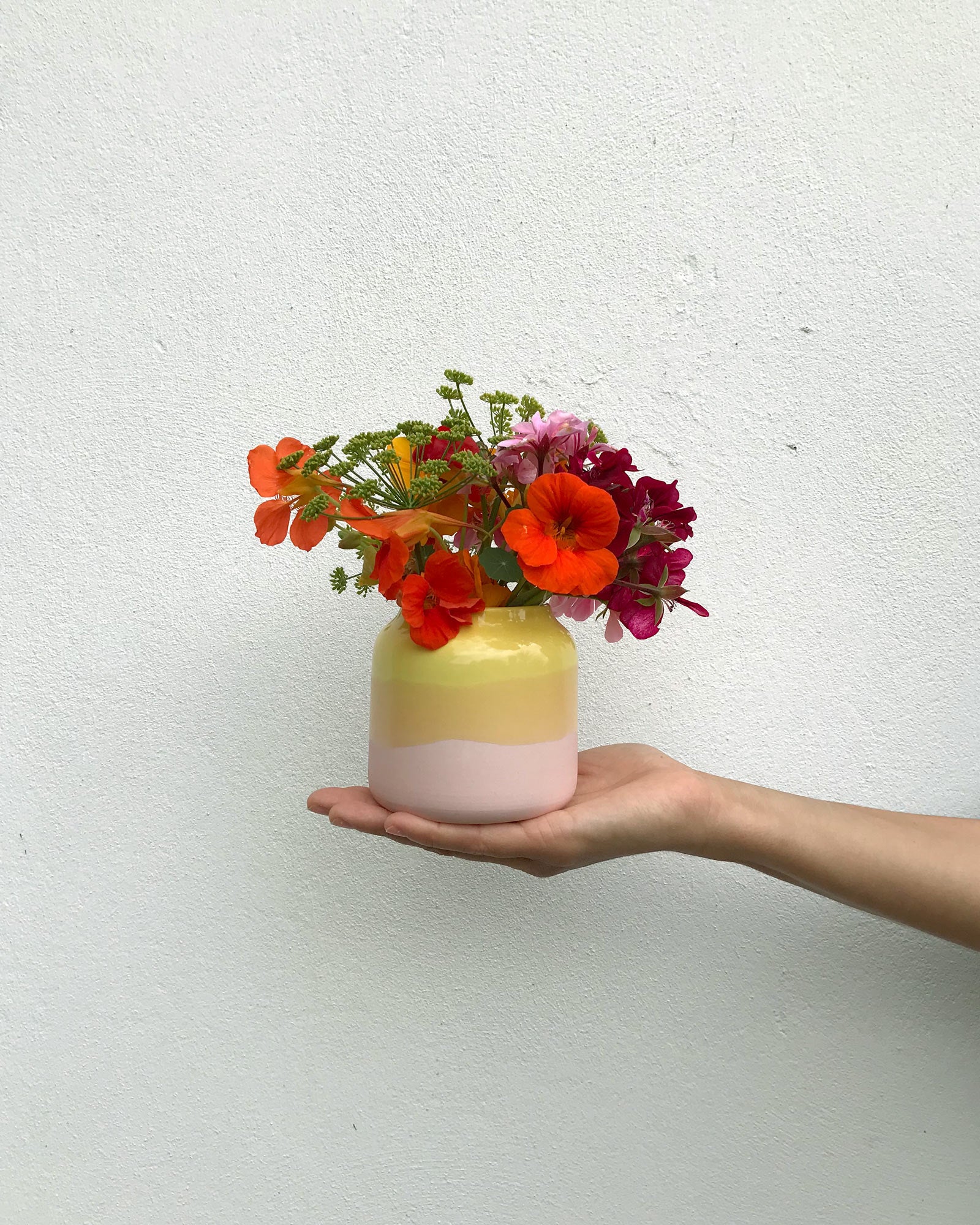 Eine Hand hält eine Vase mit farbenfrohen Blumen vor einer weißen Wand.