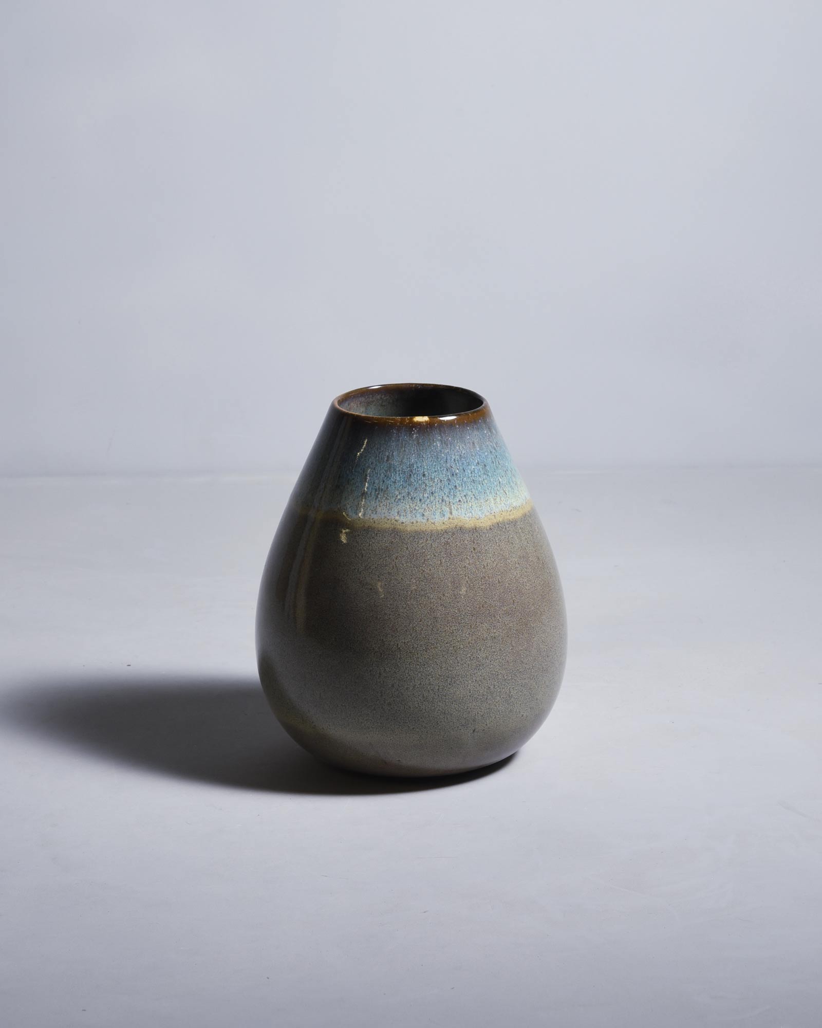 Graue Keramikvase mit blauer und brauner Glasur auf hellem Hintergrund, wirft einen Schatten nach links.