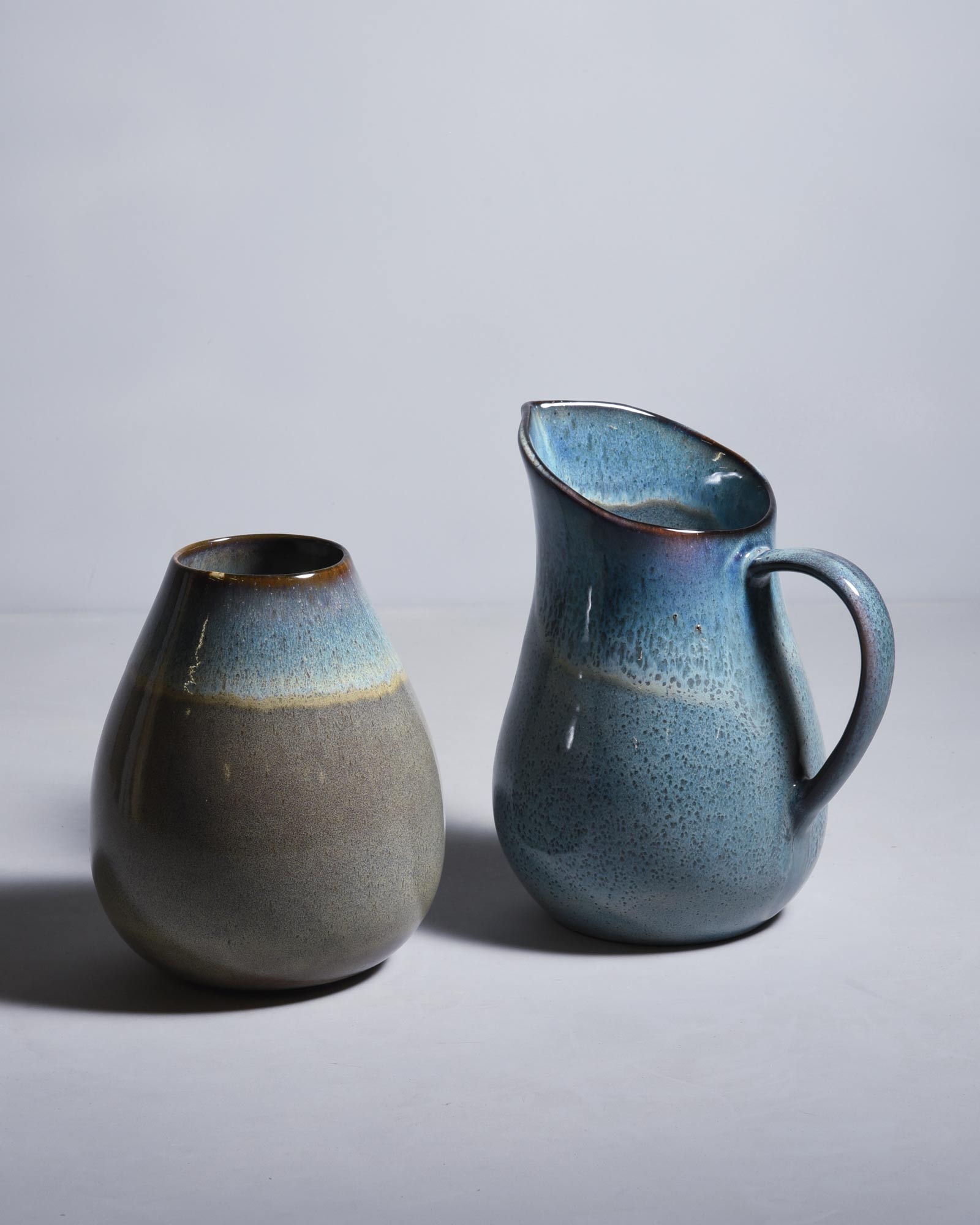 Zwei Keramikkrüge auf einem weißen Hintergrund, einer bauchig und der andere mit Henkel und Tülle, in Blau- und Brauntönen.