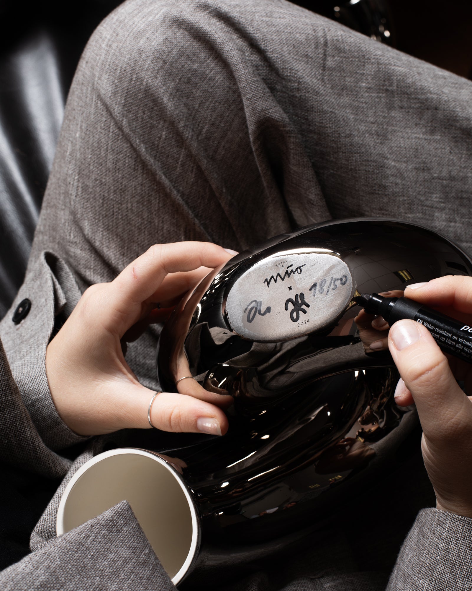 Eine Person signiert mit einem Stift den Boden einer glänzenden, schwarzen Vase, die auf ihrem Schoß gehalten wird.