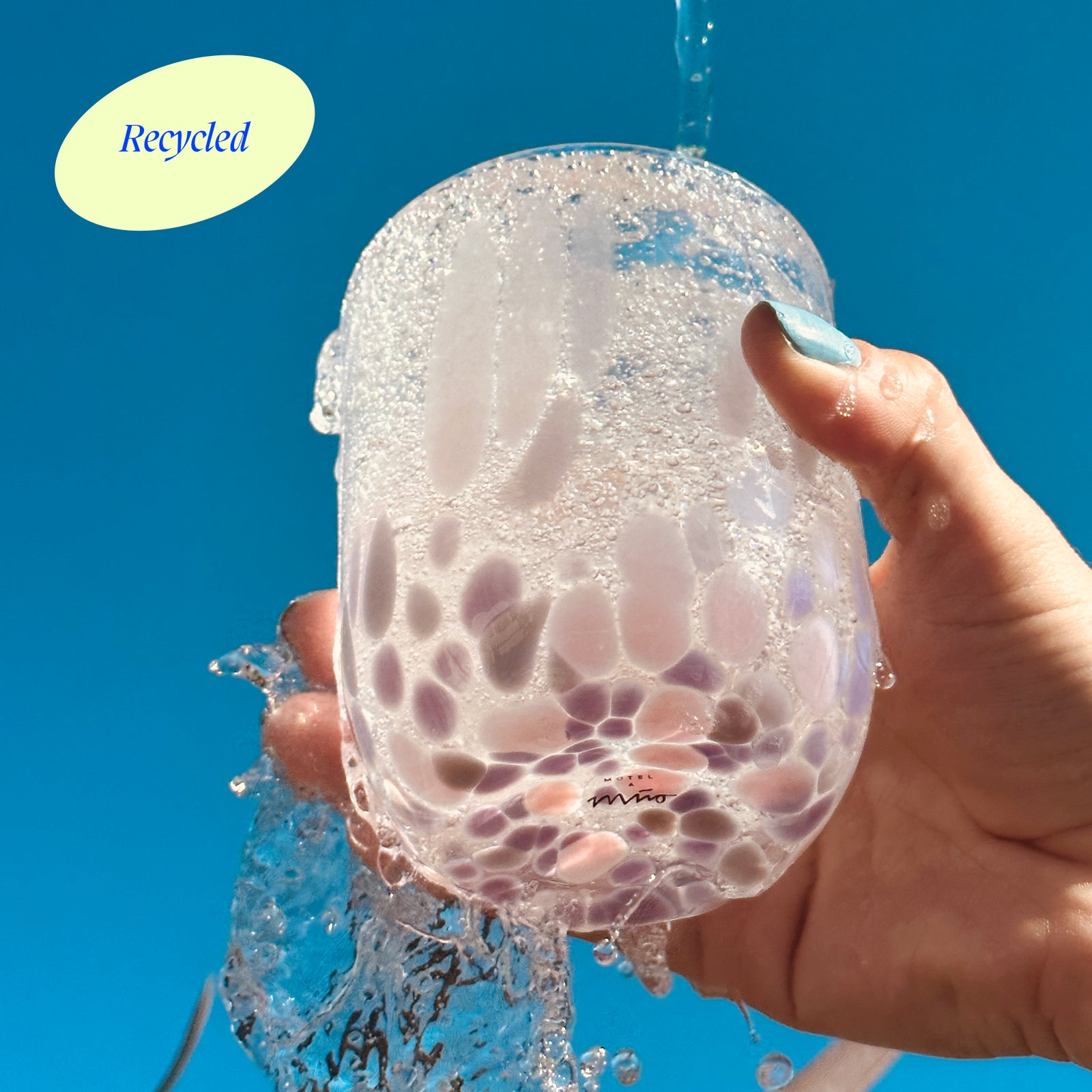 Eine Hand hält ein transparentes, gepunktetes Glas mit Wasser vor blauem Himmel; Text "Recycled" oben links.