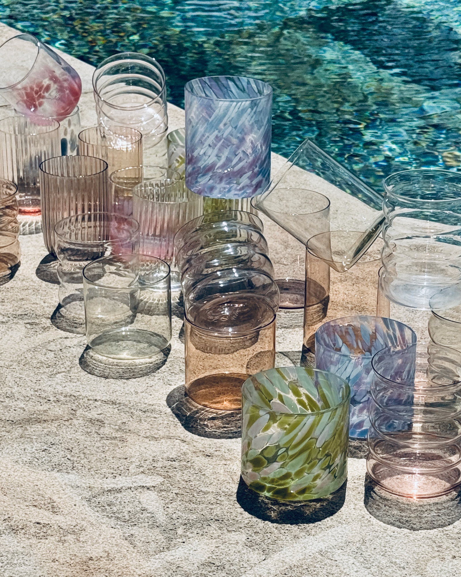 Mehrere bunte, gemusterte Glasbecher stehen auf einem Steintisch neben einem Pool in der Sonne.