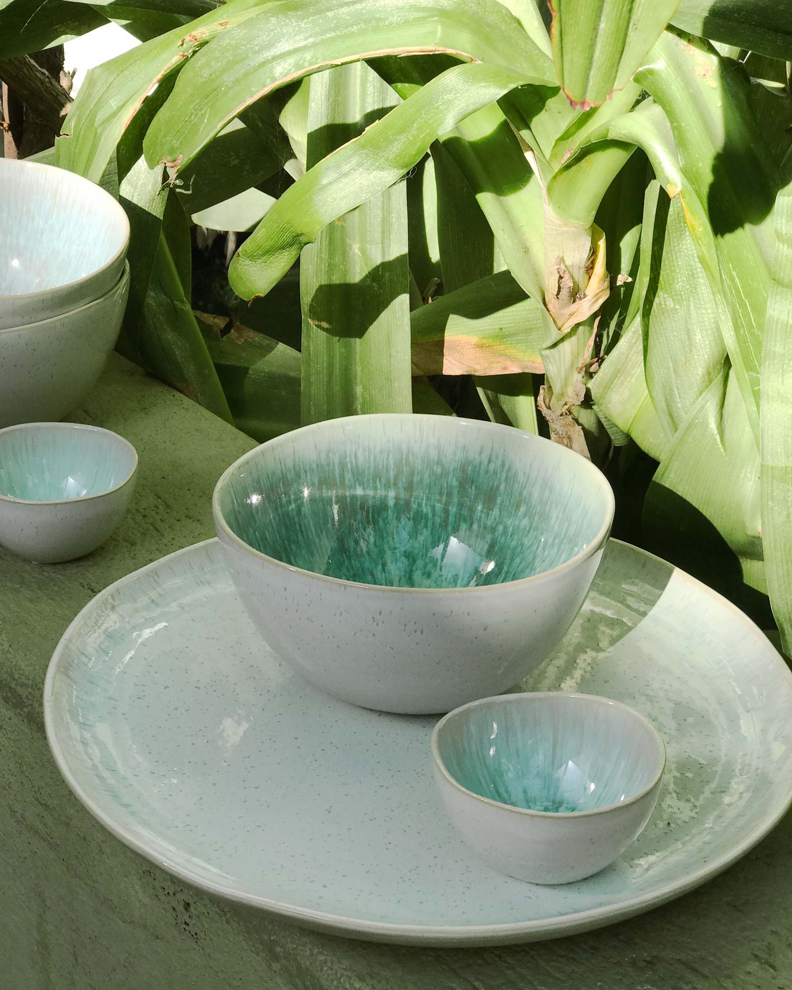 Keramikschalen in verschiedenen Größen mit grün-blauer Glasur, arrangiert auf einem Teller vor grünen Blättern.