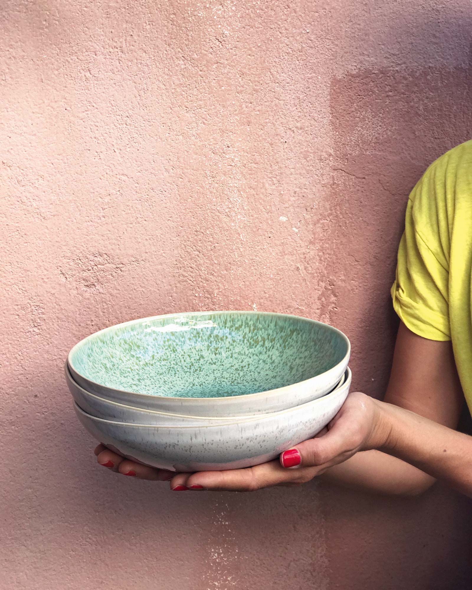 Eine Person hält gestapelte Keramikschalen mit grünem Muster vor einer rosa Wand.