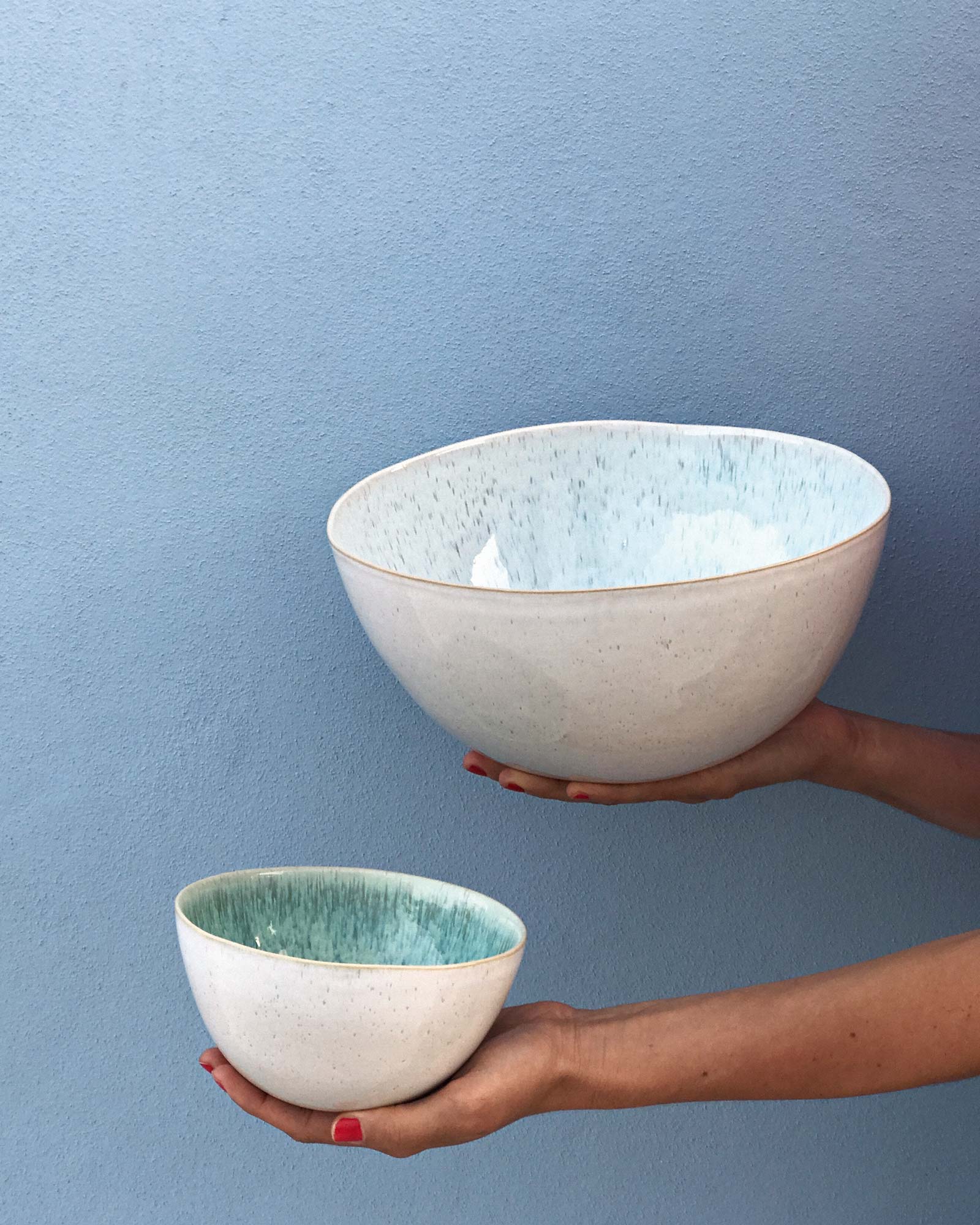 Zwei Keramikschüsseln, eine größer, eine kleiner, werden vor einer hellblauen Wand in Händen gehalten.