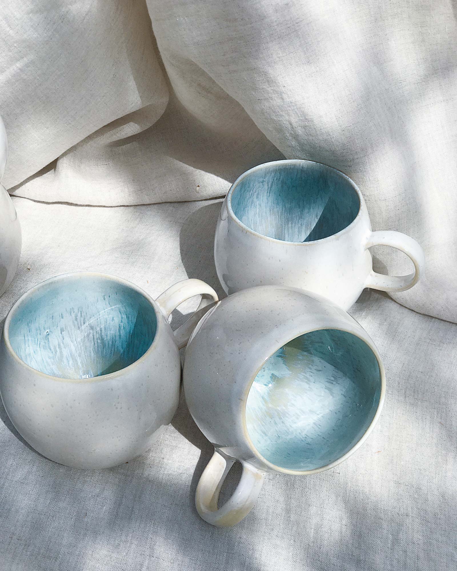 Drei weiße Tassen mit blauem Inneren auf einem hellen Stoff drapiert.