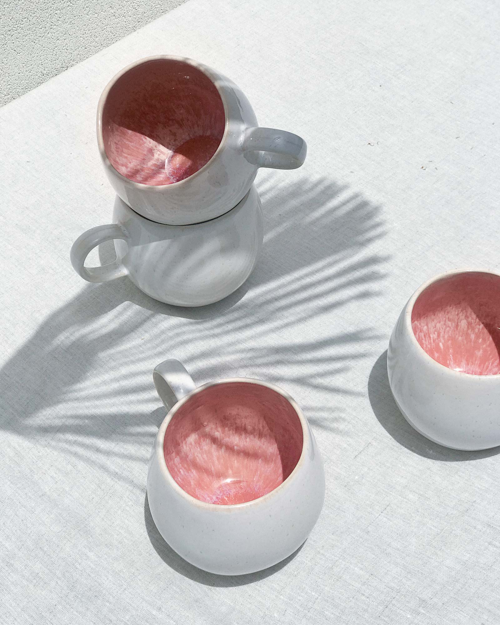 Vier weiße Tassen mit roter Innenseite auf einem weißen Tuch, mit Schatten von Palmblättern.