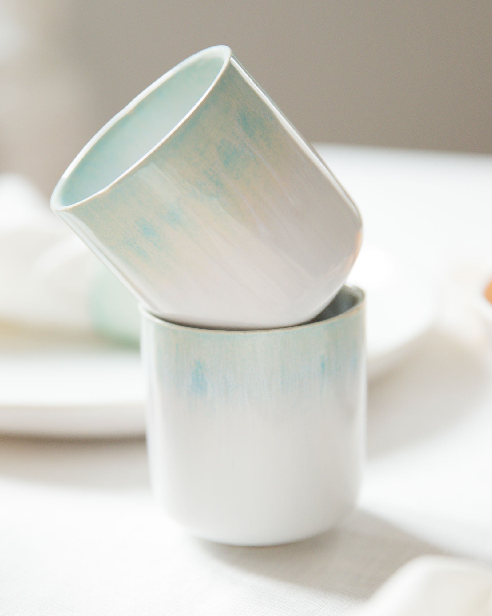 Zwei ineinander gestapelte, pastellfarbene Tassen auf einem Tisch mit weißem Tischtuch.