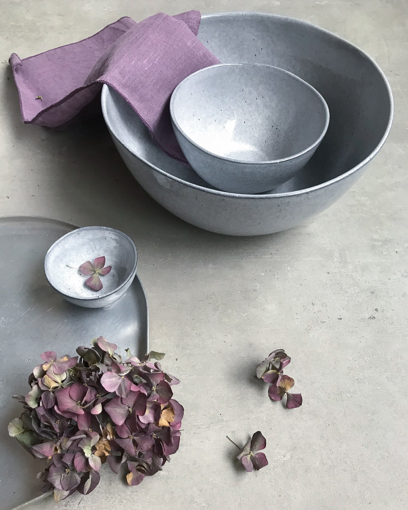 Drei graue Schalen, eine lila Serviette und getrocknete Blumen auf einem grauen Tisch.