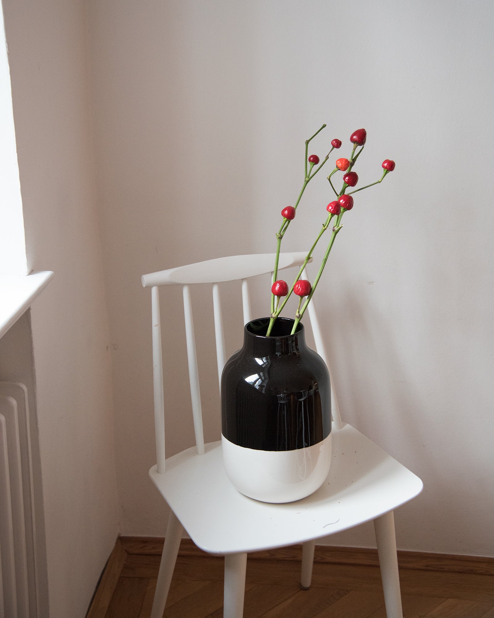Eine schwarz-weiße Vase mit roten Beerenzweigen steht auf einem weißen Stuhl vor einer weißen Wand.