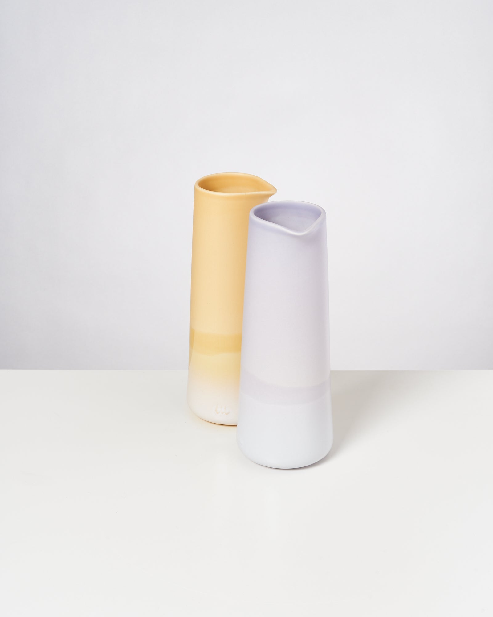 Zwei minimalistische, pastellfarbene Krüge, einer gelb, der andere lila, stehen auf einem weißen Tisch vor hellem Hintergrund.