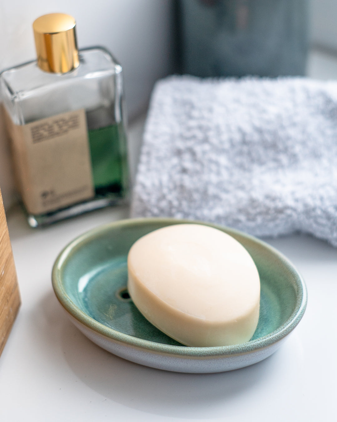 Ein Stück Seife liegt auf einer grünen Seifenschale, daneben eine Flasche Parfüm und ein graues Handtuch.