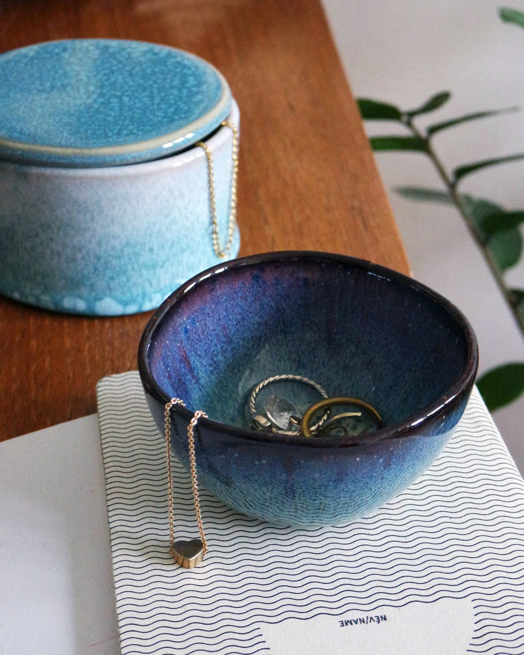Eine blaue Keramikschale mit Schmuckstücken steht auf einem Buch, daneben eine passende Dose auf einem Holztisch.