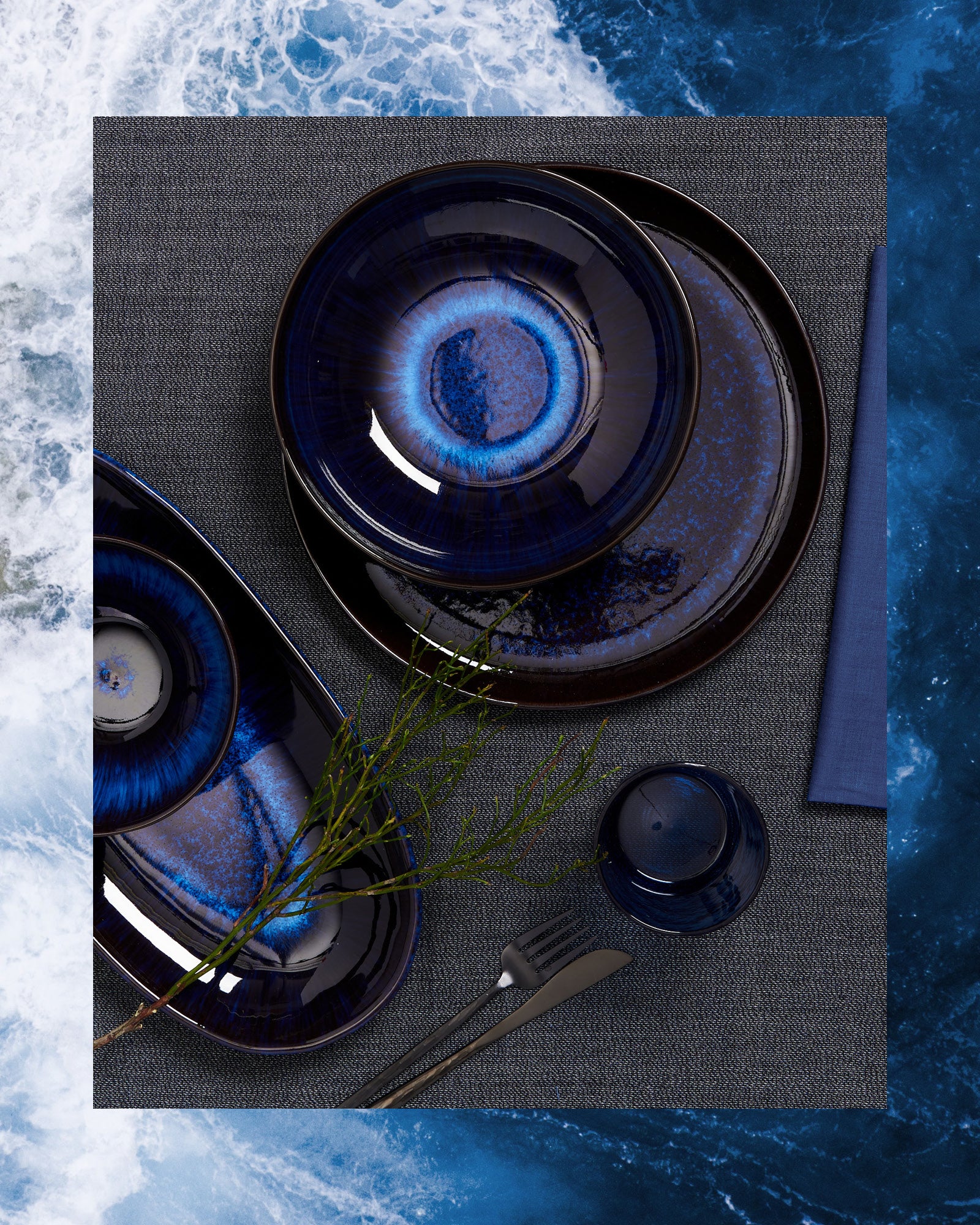 Teller und Schalen in dunklem Blau mit hellerem Zentrum auf grauem Tischtuch, dekoriert mit einem grünen Zweig.