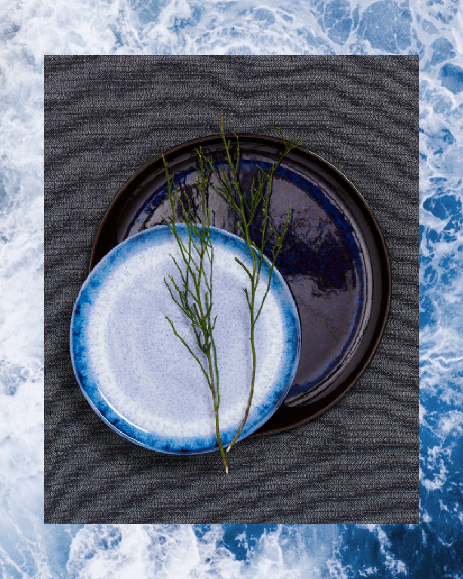 Zwei Keramikteller, einer blau-weiß, der andere dunkelblau, liegen auf grauem Stoff, verziert mit Zweigen.