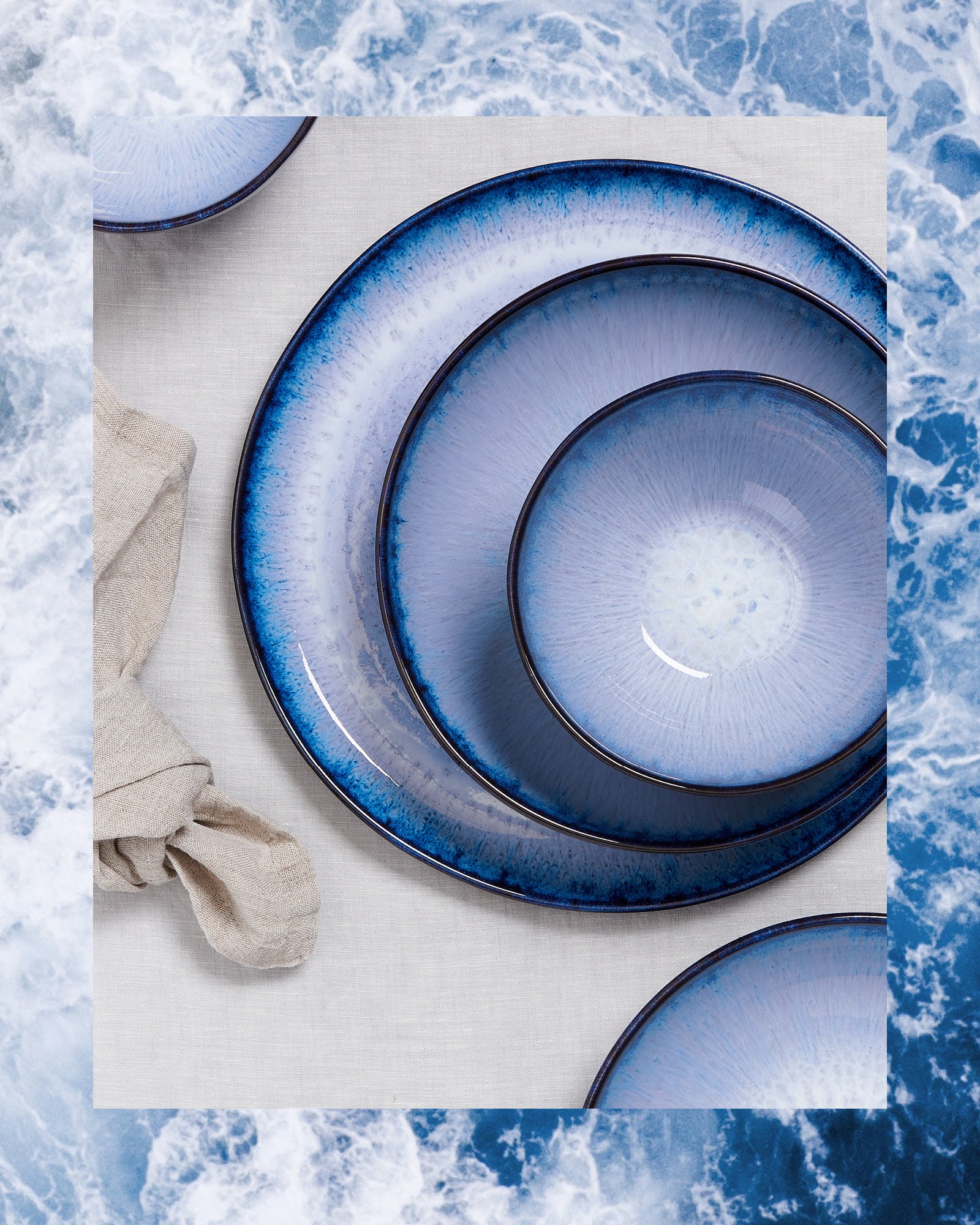 Blau-weiße Teller in verschiedenen Größen auf einem hellen Tuch, umgeben von einem Wellenmuster.