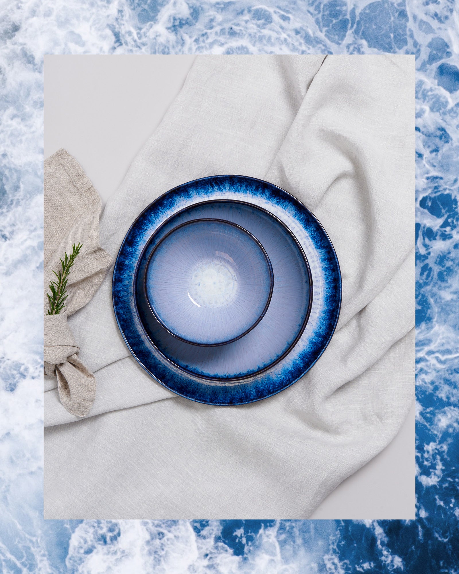 Zwei blaue Teller auf weißem Stoff, umgeben von einem Rahmen aus Wellenmuster, neben einem Stoffservietten-Bündel.