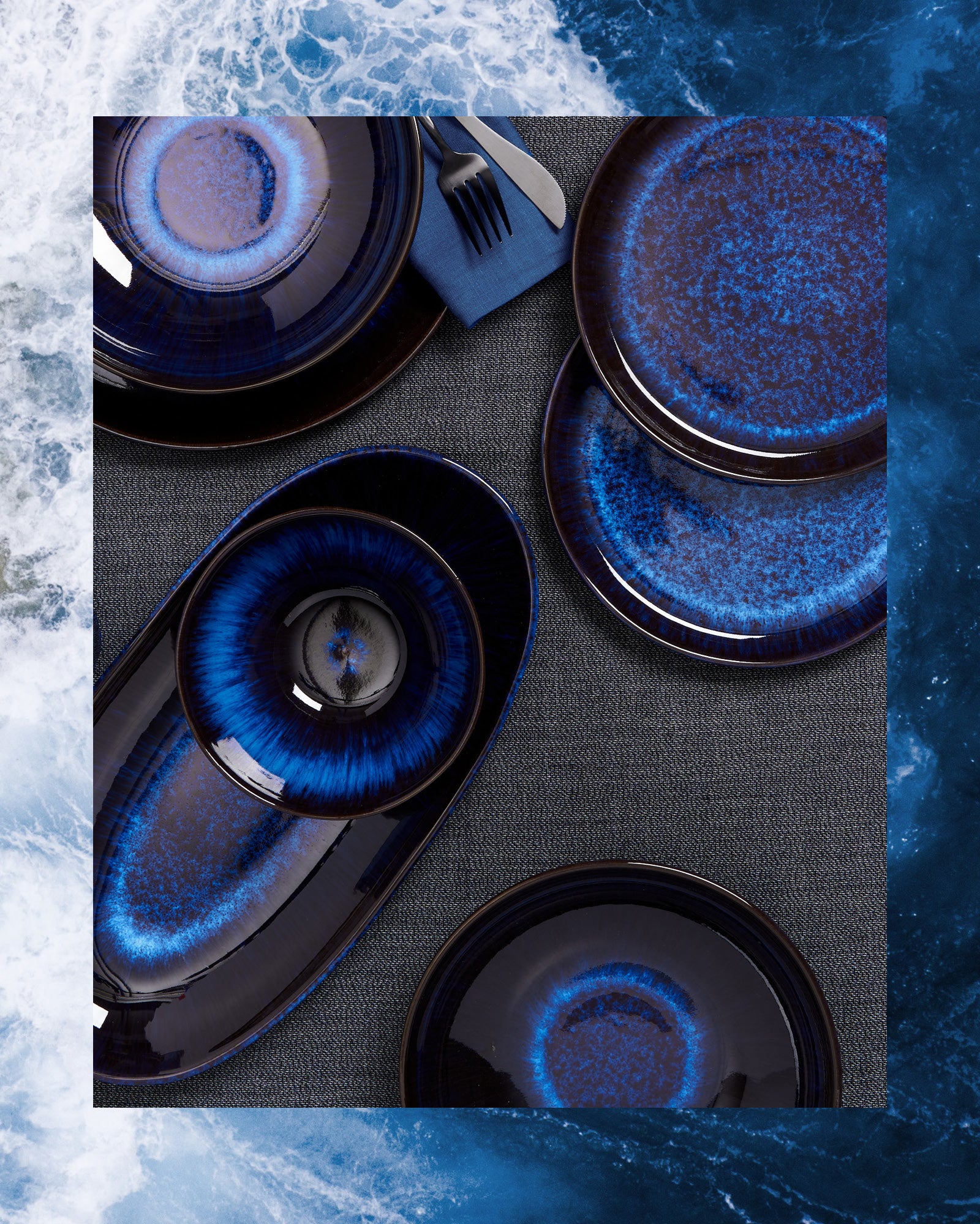 Verschiedene dunkelblaue Keramikteller und Besteck auf einem grauen Tischtuch mit einem maritimen Hintergrund.