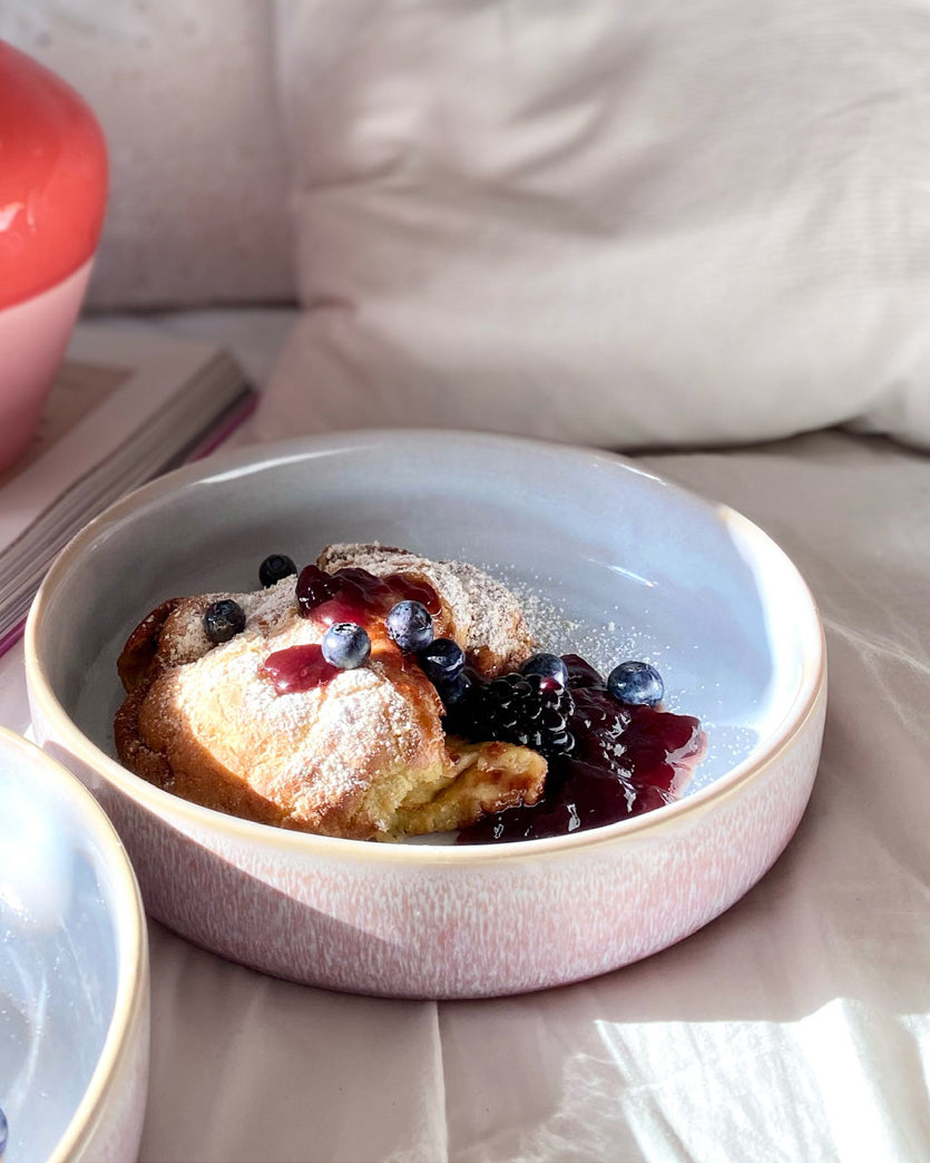 Eine Schüssel mit Pfannkuchen, garniert mit Puderzucker, Blaubeeren und Brombeeren, auf einem Tisch bei Sonnenlicht.