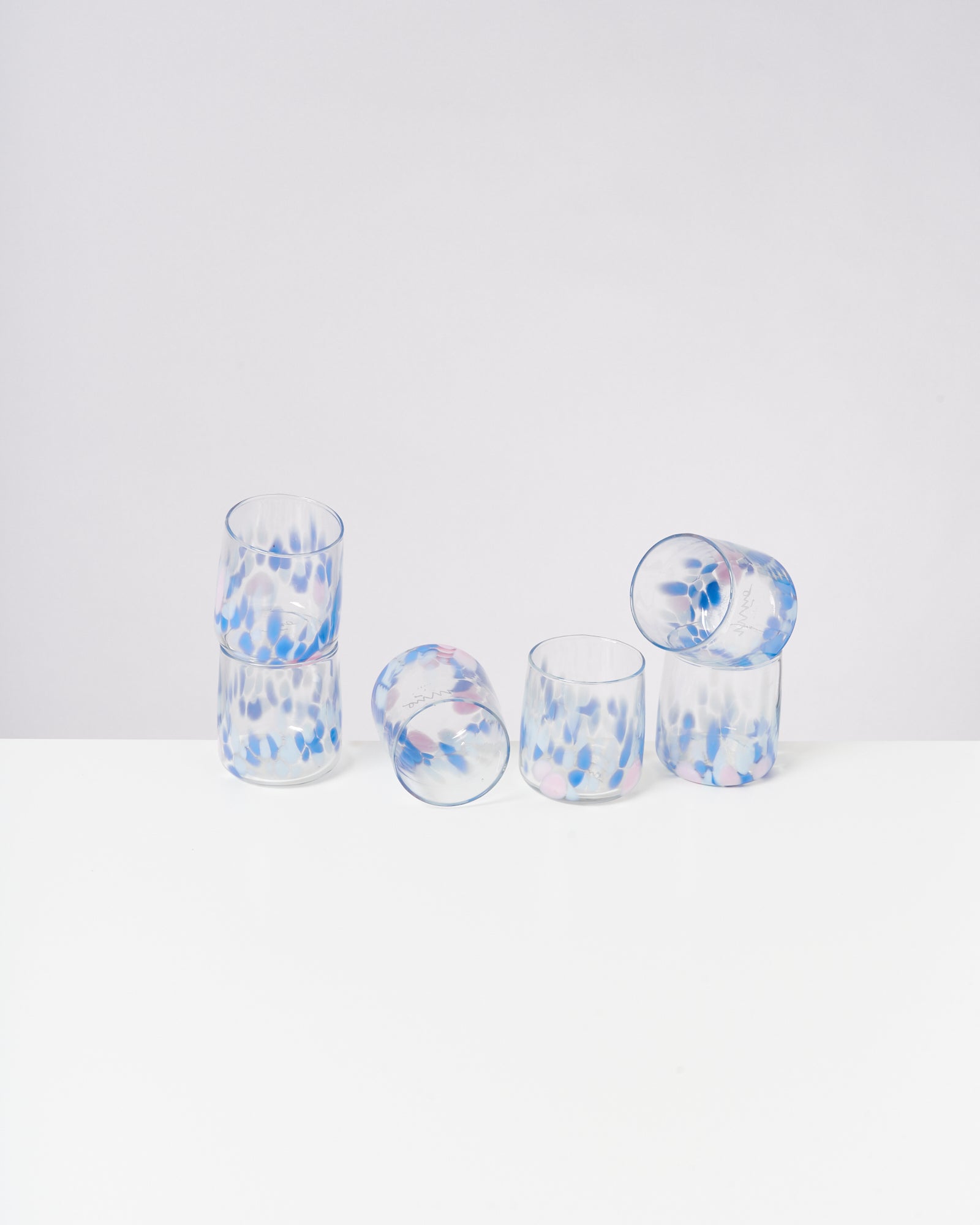 Sechs Gläser mit blauen und rosa Punkten stehen und liegen auf einem weißen Tisch vor einem weißen Hintergrund.