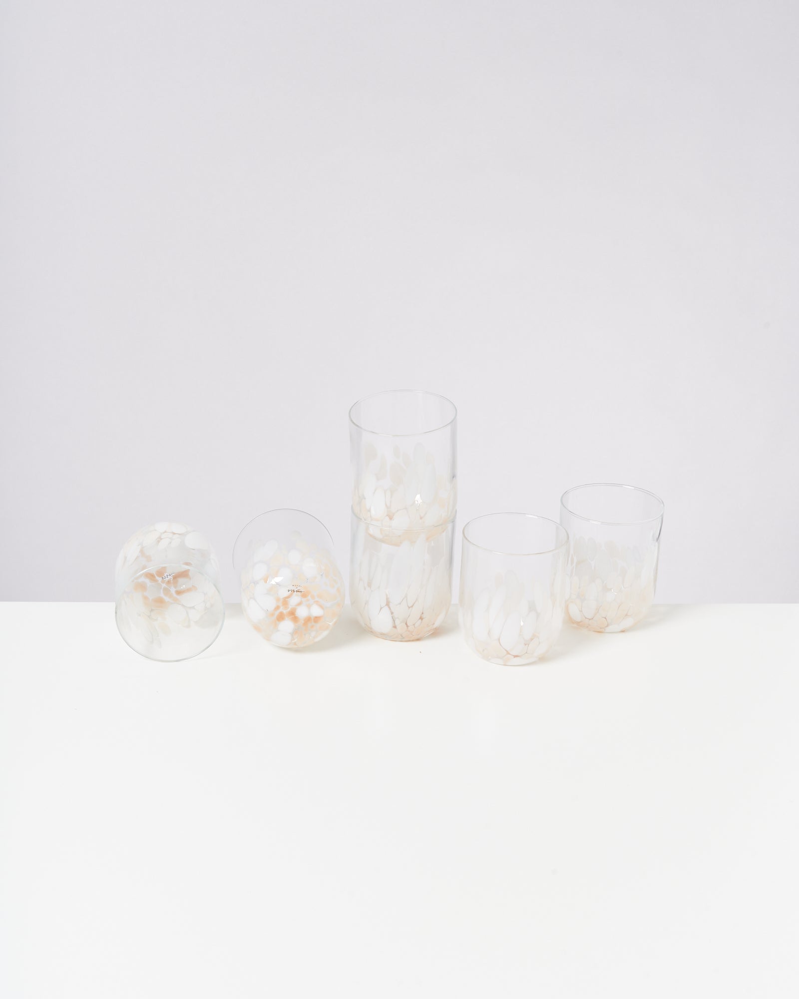 Fünf gläserne Becher mit weißem, fleckigem Muster stehen auf einem weißen Tisch vor einem grauen Hintergrund.