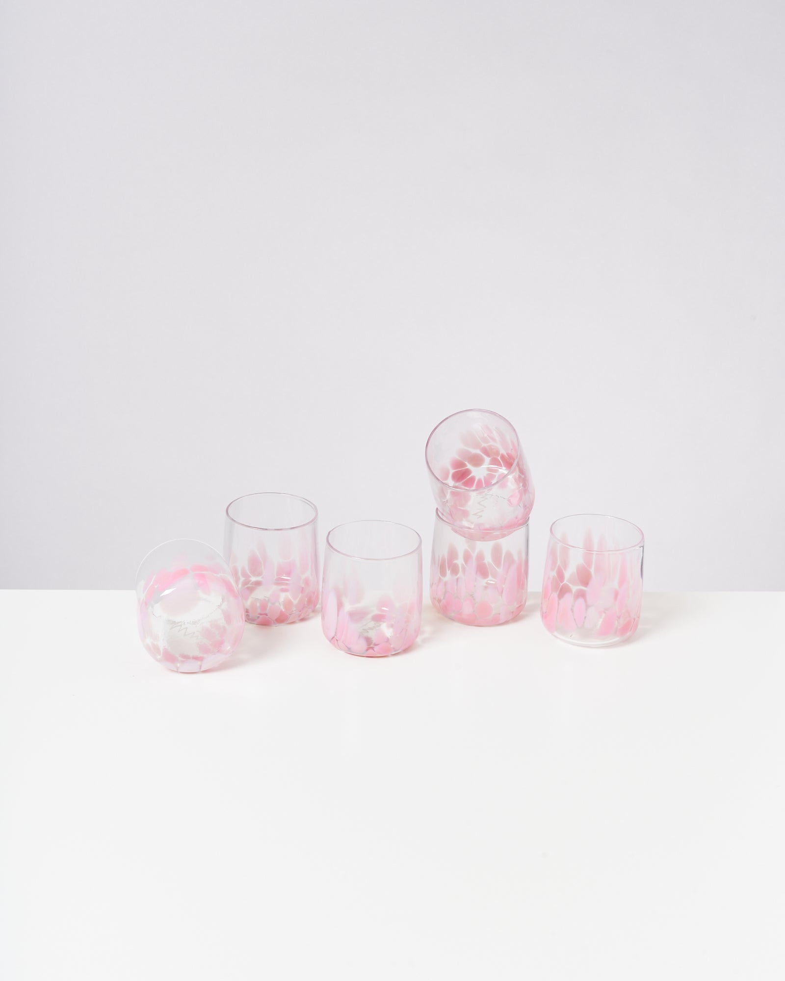 Fünf Glasbecher mit rosa und weißen Mustern auf einem weißen Tisch vor einem hellgrauen Hintergrund.