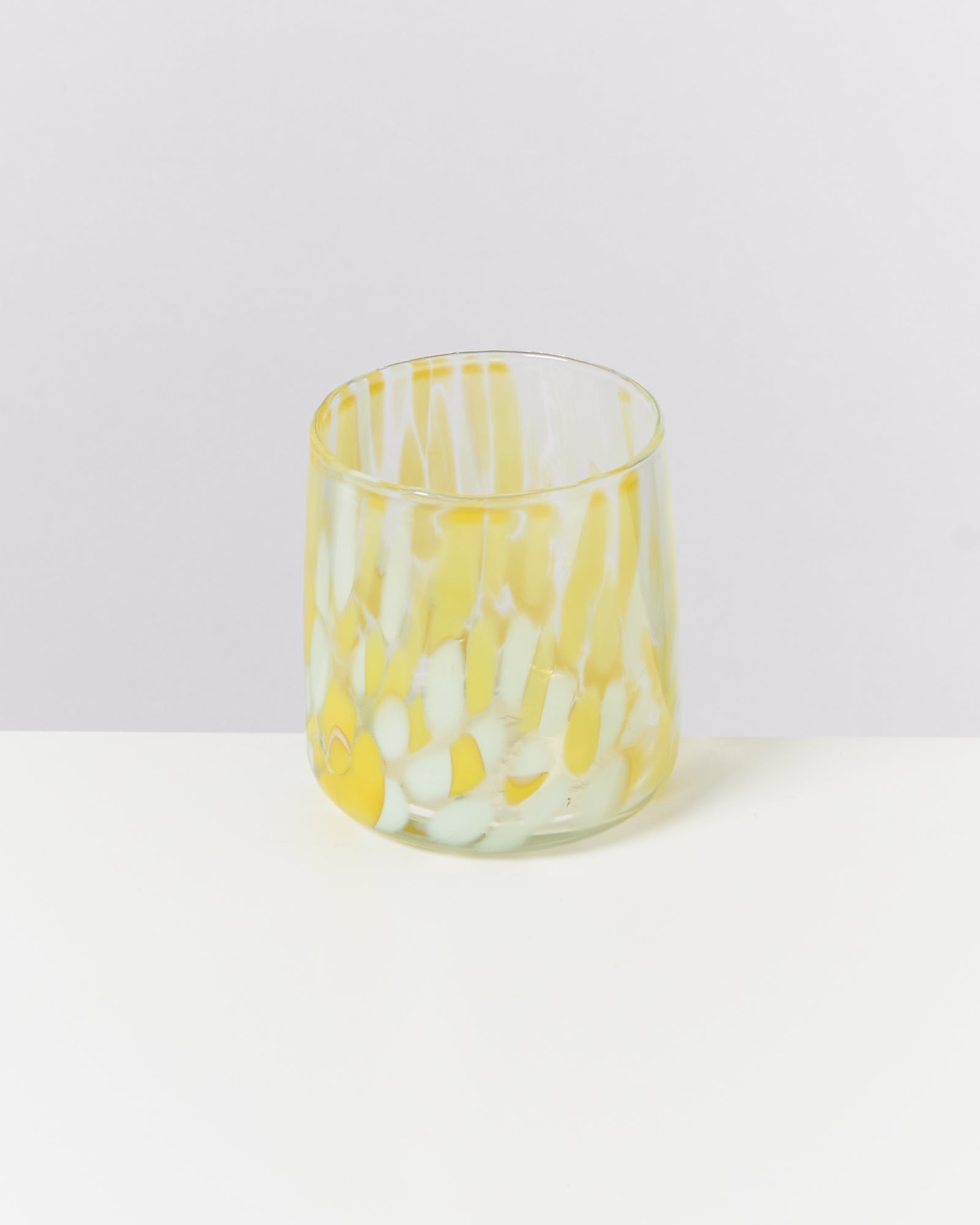Ein buntes Glas mit gelben und weißen Mustern steht auf einer weißen Oberfläche vor einem grauen Hintergrund.