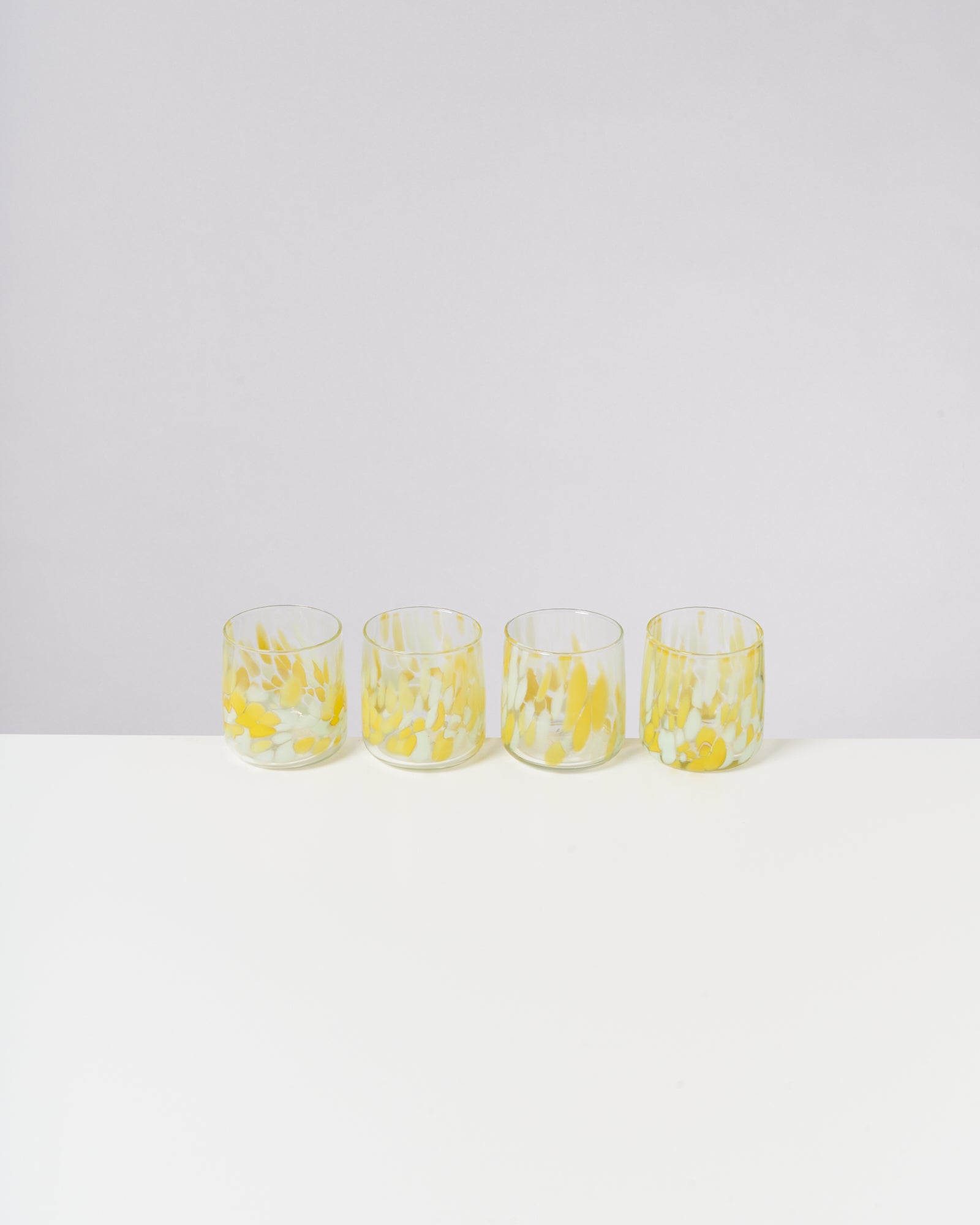 Vier bunte Gläser mit gelben und weißen Mustern stehen in einer Reihe auf einem weißen Tisch vor grauem Hintergrund.
