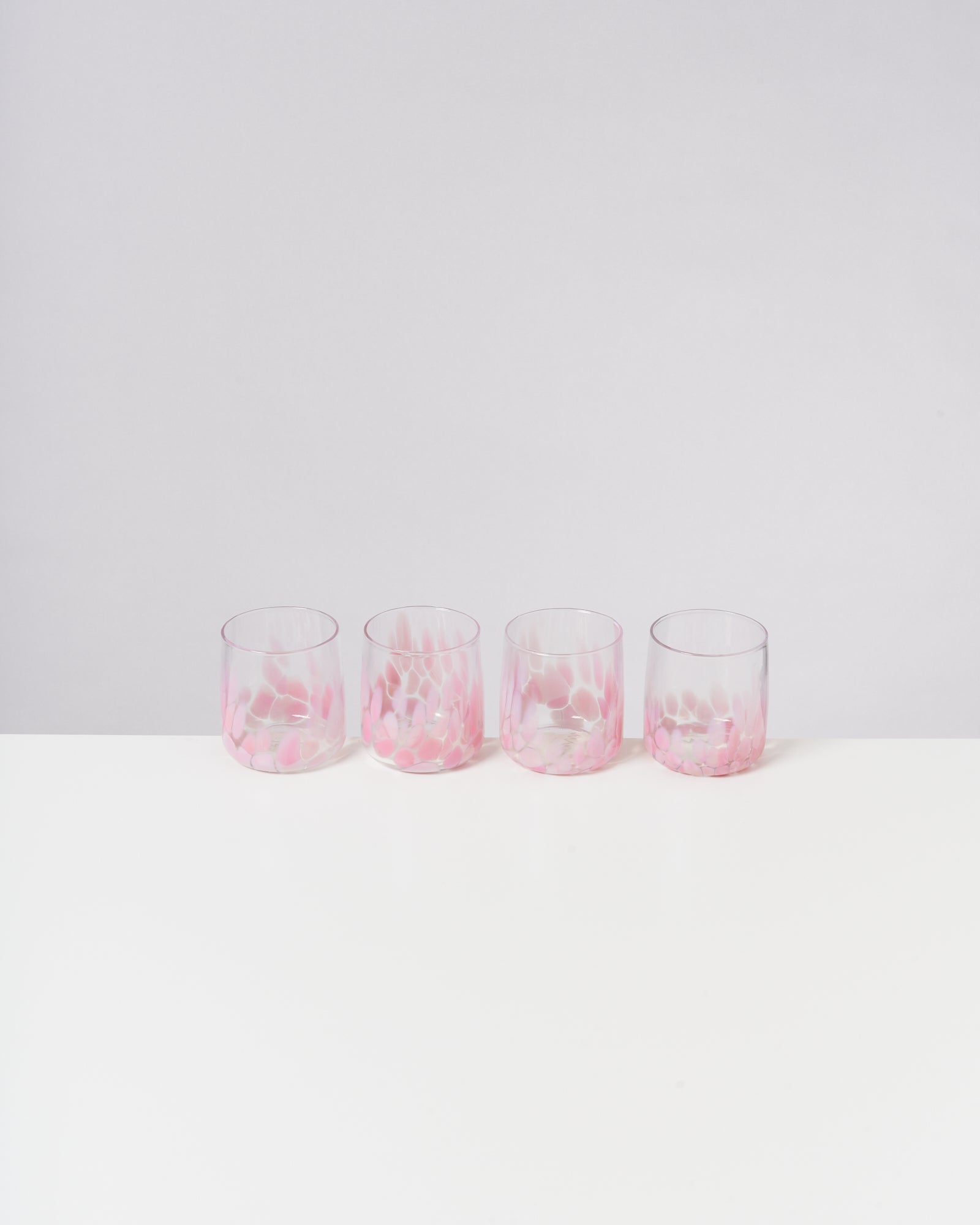 Vier Gläser mit rosa Muster stehen in einer Reihe auf einem weißen Tisch vor hellem Hintergrund.