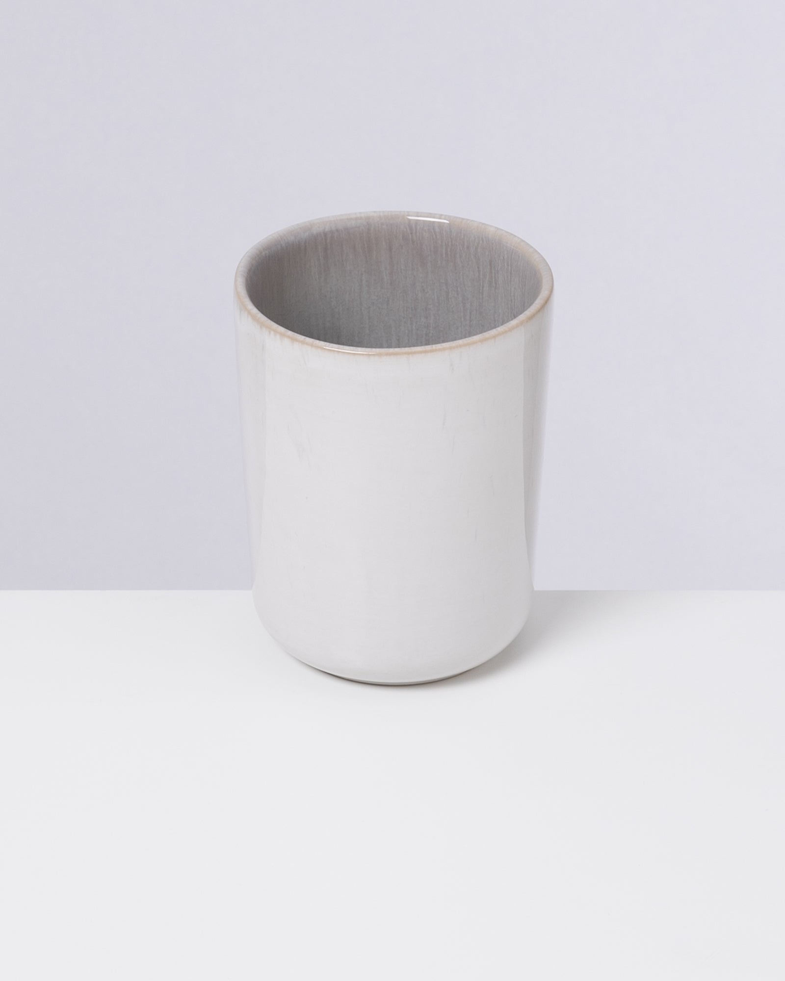 Grauer Keramikbecher steht auf einem weißen Untergrund vor einem hellgrauen Hintergrund.