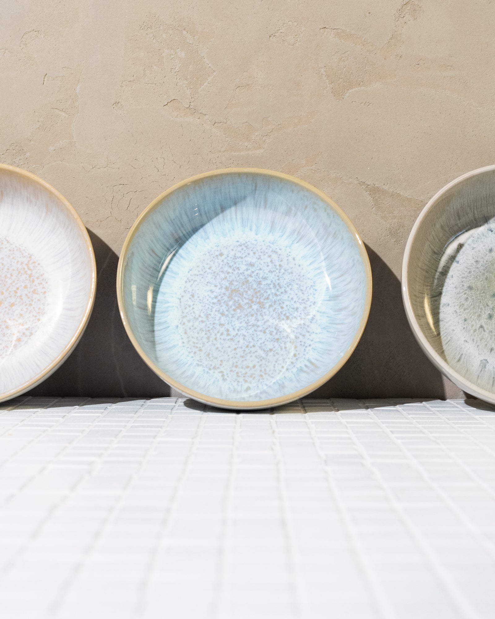 Drei Keramikteller in verschiedenen Farben auf hellem Mosaikboden vor einer beigen Wand angeordnet.