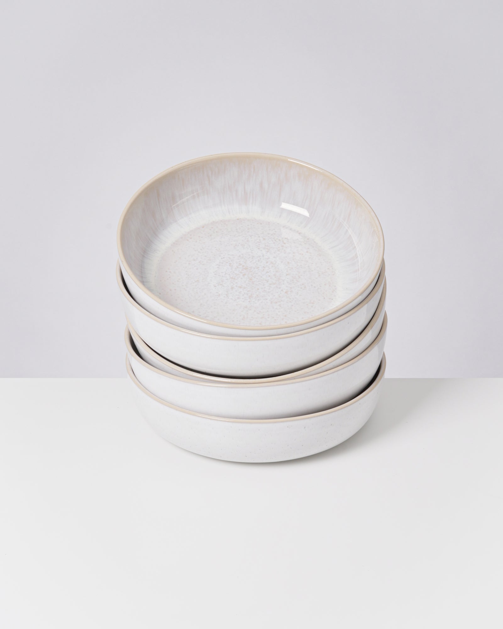 Ein Stapel aus vier weißen Keramikschalen mit leichtem Muster steht auf einem weißen Tisch vor einem grauen Hintergrund.