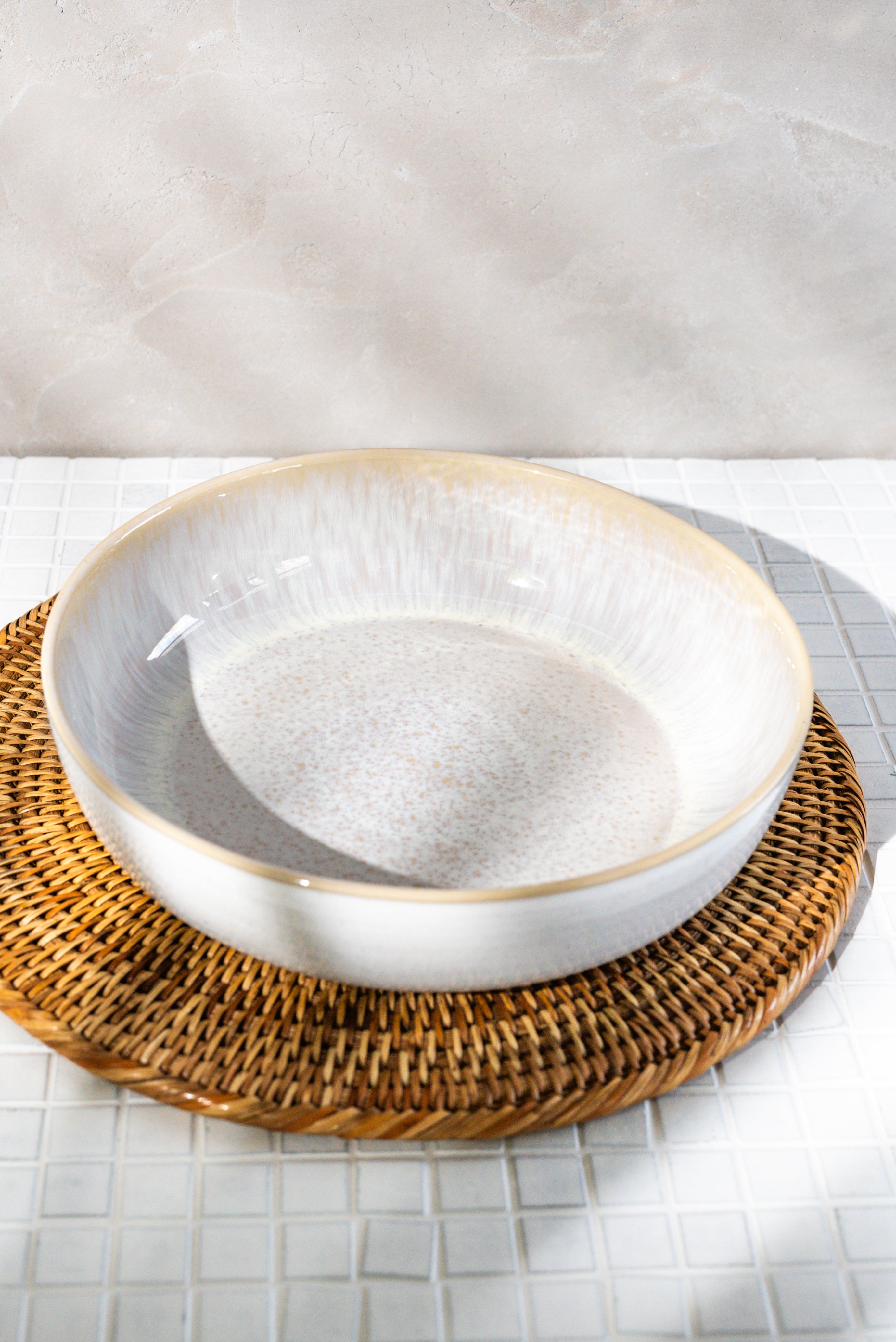 Große, beige Keramikschale auf rundem, geflochtenem, braunem Tischset auf weißen Fliesen.