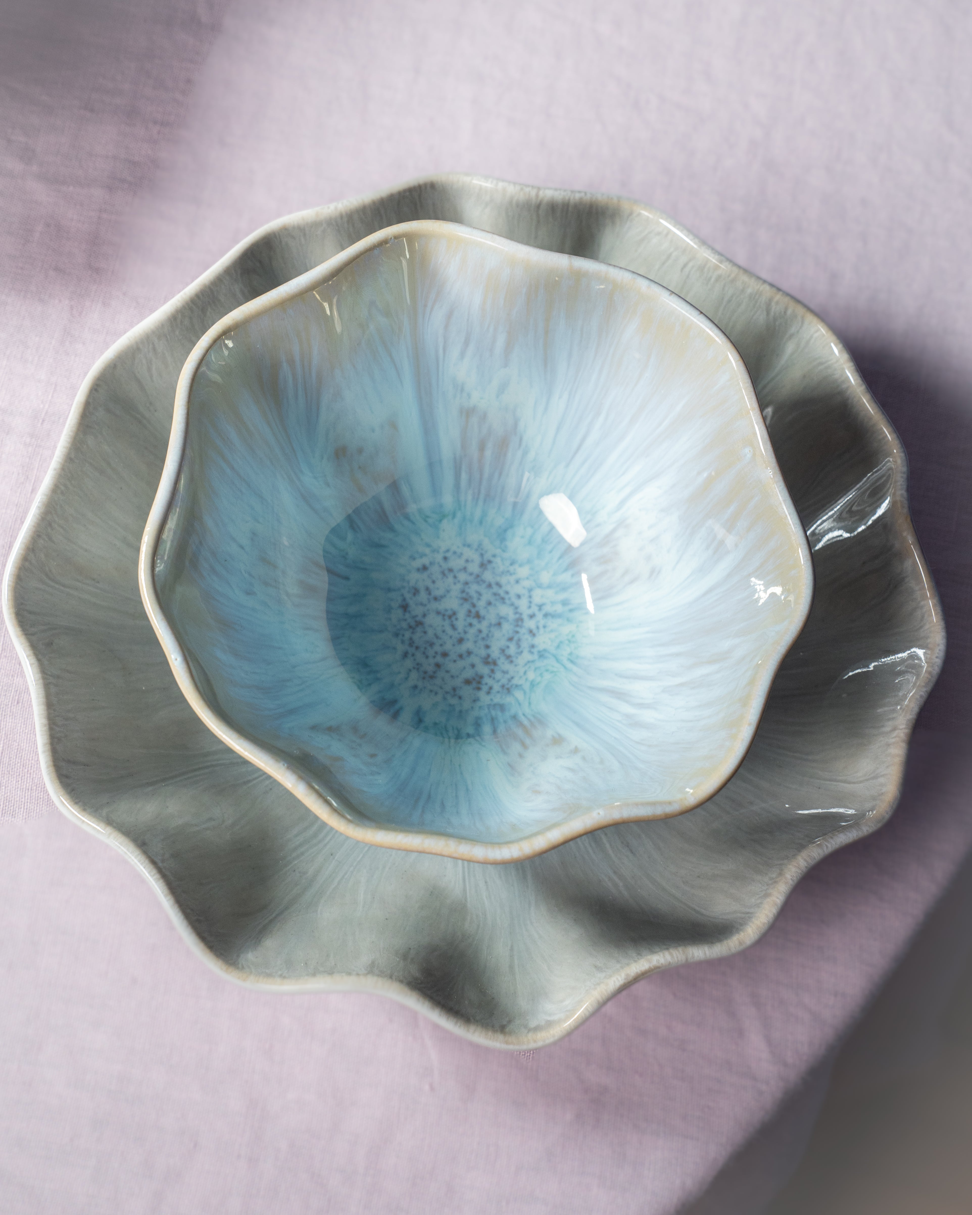 Zwei übereinander gestapelte, blütenförmige Keramikschalen in Blau- und Grautönen auf einem rosa Stoffhintergrund.