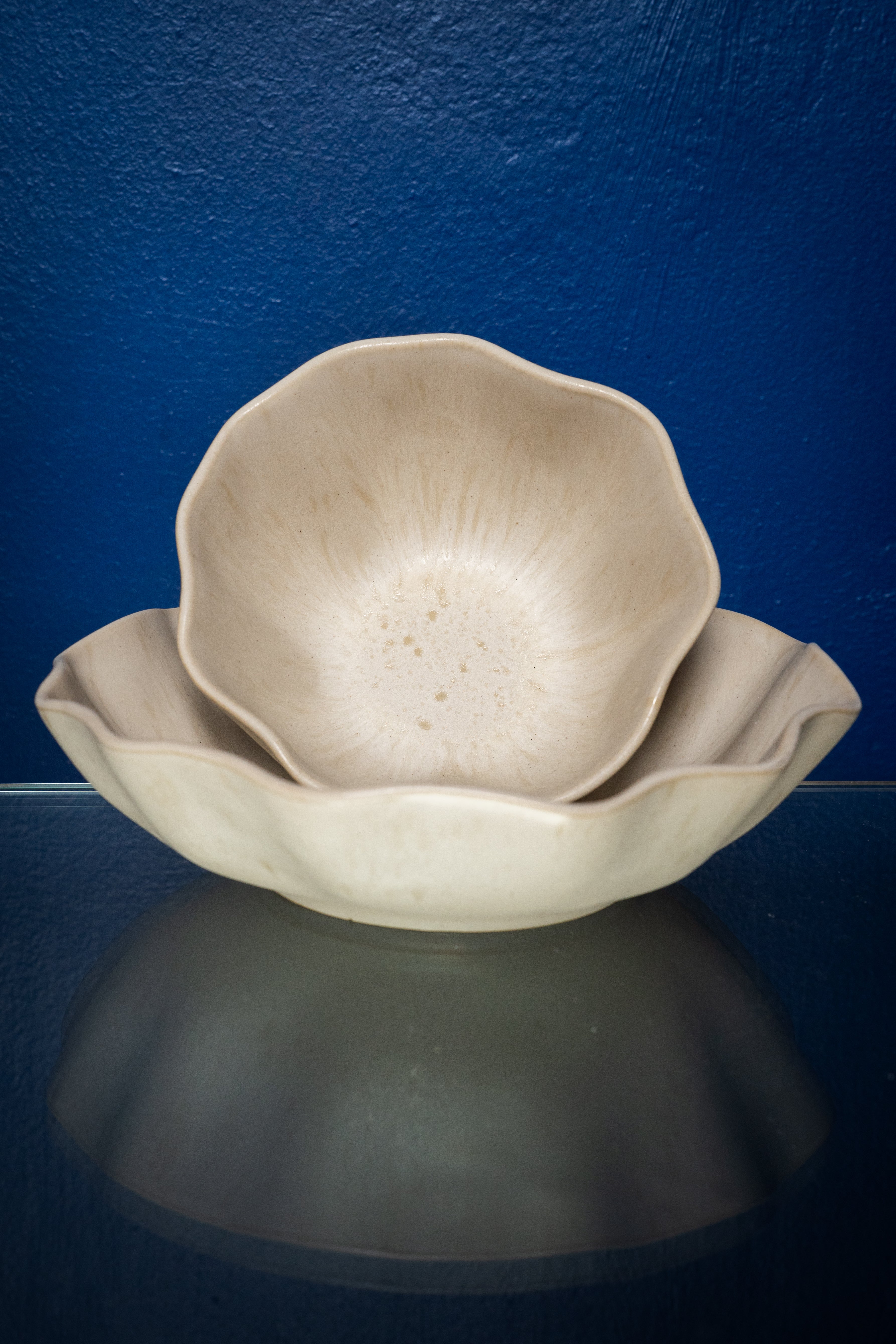 Zwei beige, blütenförmige Schalen stehen ineinander vor einer blauen Wand auf einem Glasoberfläche.