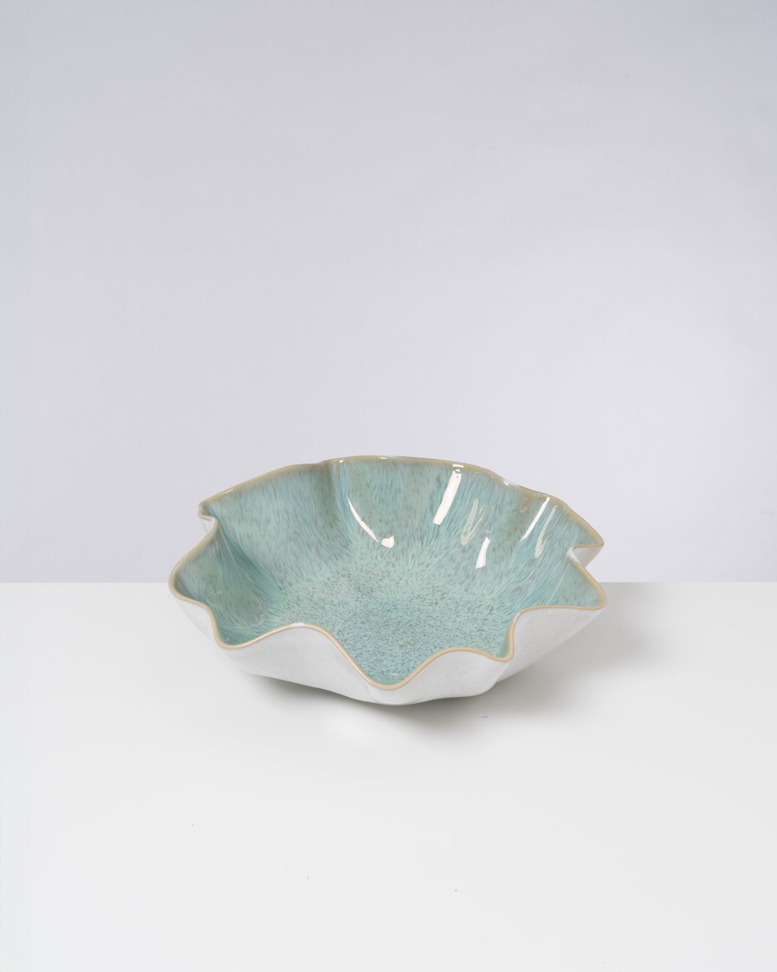 Eine dekorative Schale mit welligem Rand, blau-grüner Innenglasur und weißer Außenseite auf weißem Hintergrund.
