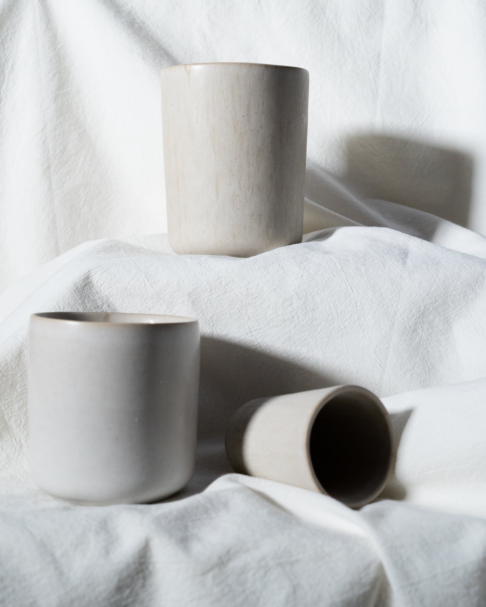 Drei schlichte, helle Keramiktassen auf weißem, gefaltetem Stoff arrangiert, eine davon liegt auf der Seite.