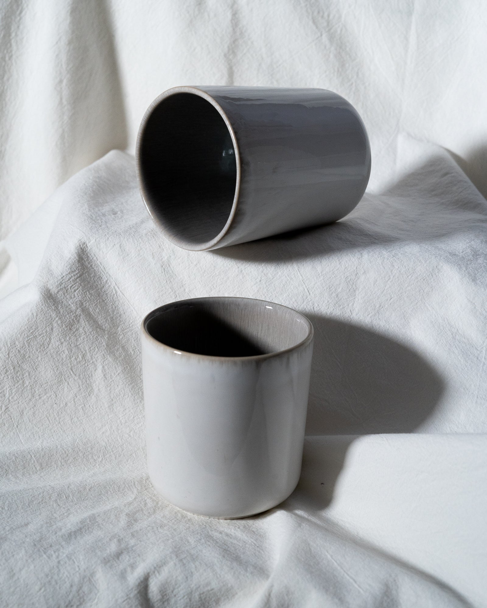 Zwei graue Keramiktassen liegen auf einem weißen, texturierten Stoffuntergrund, eine steht aufrecht, die andere liegt seitlich.