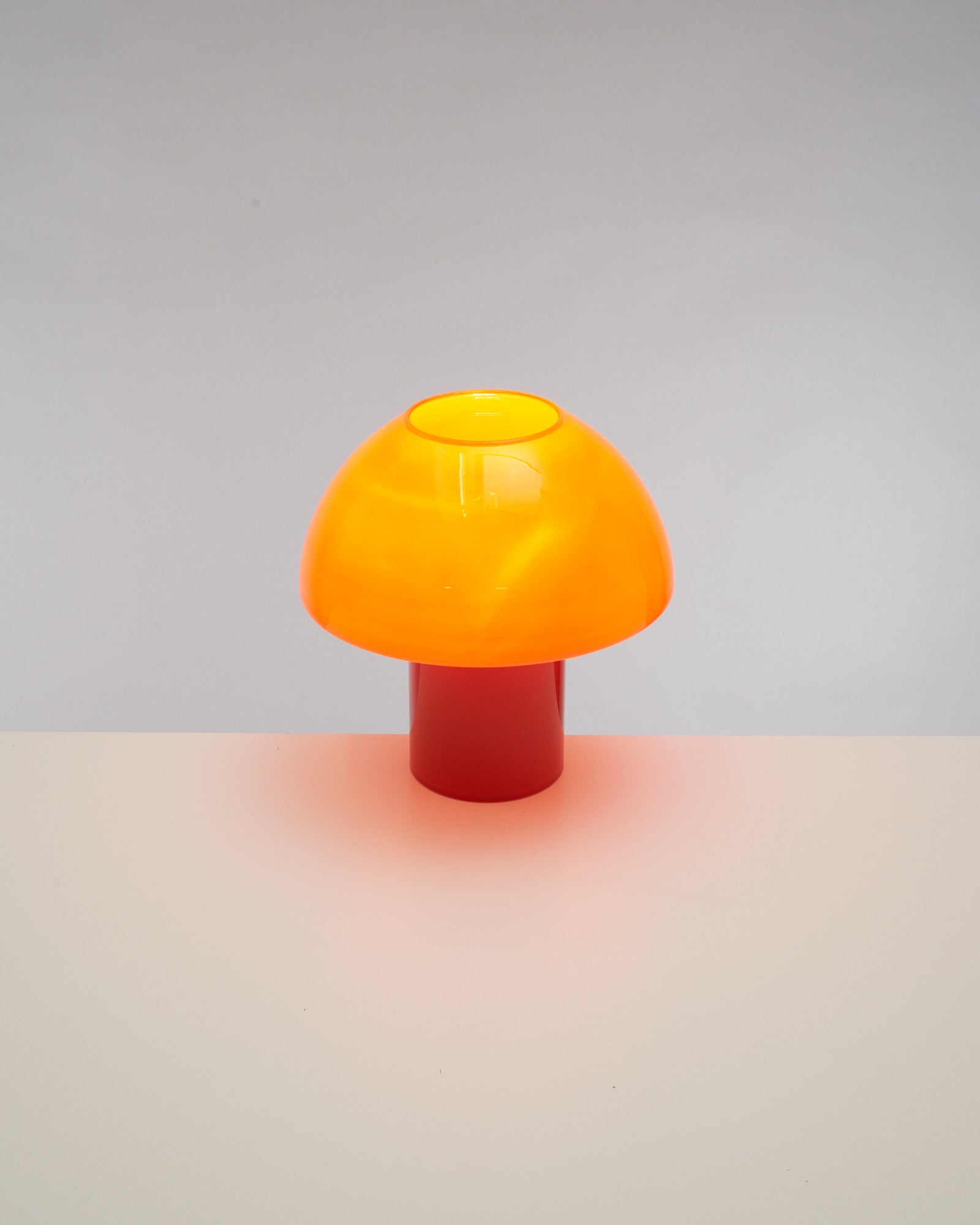 Orangefarbene Pilzlampe auf rotem Sockel vor grauem Hintergrund.