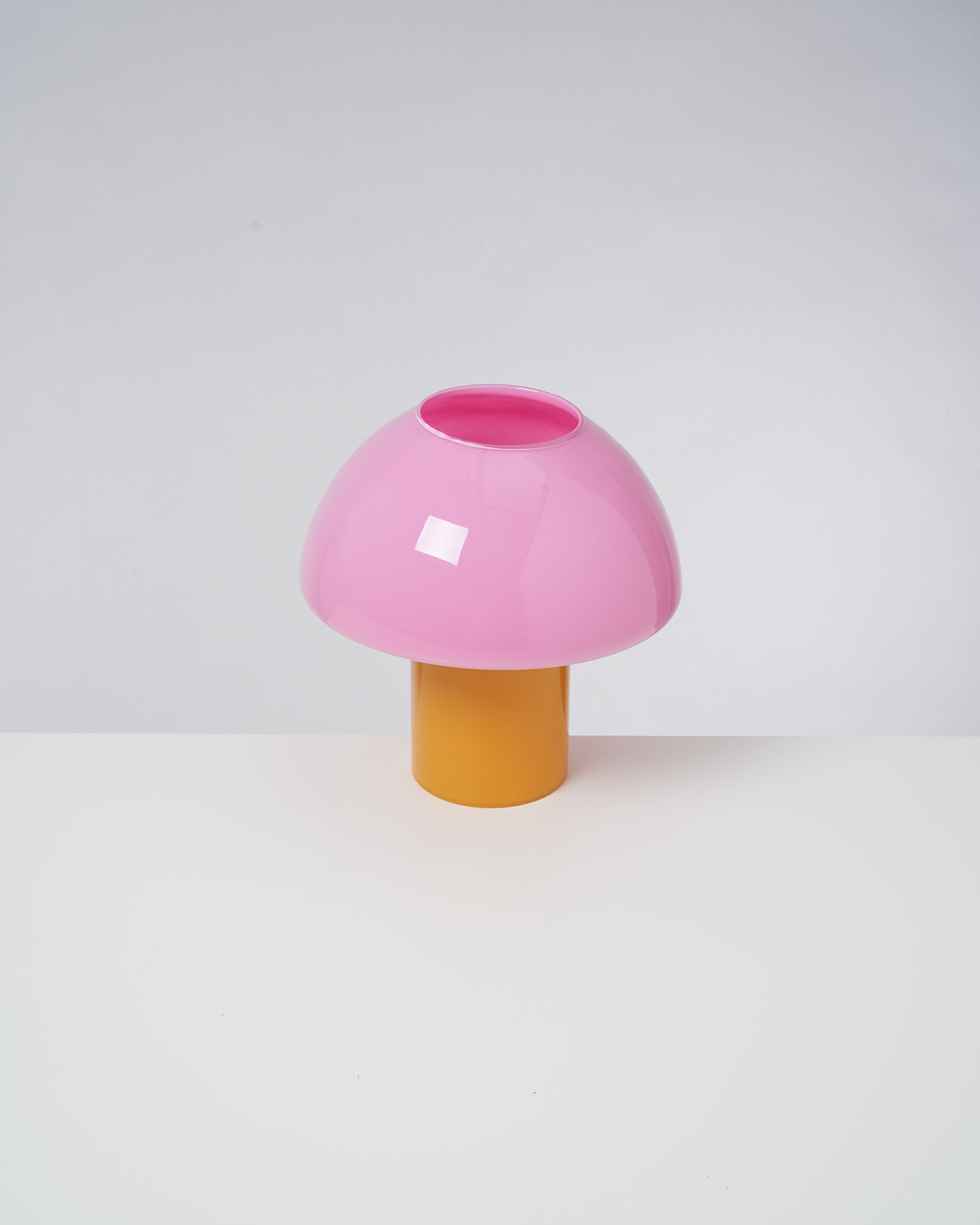 Rosa Tischlampe mit einem orangefarbenen Fuß auf einem weißen Untergrund vor grauem Hintergrund.