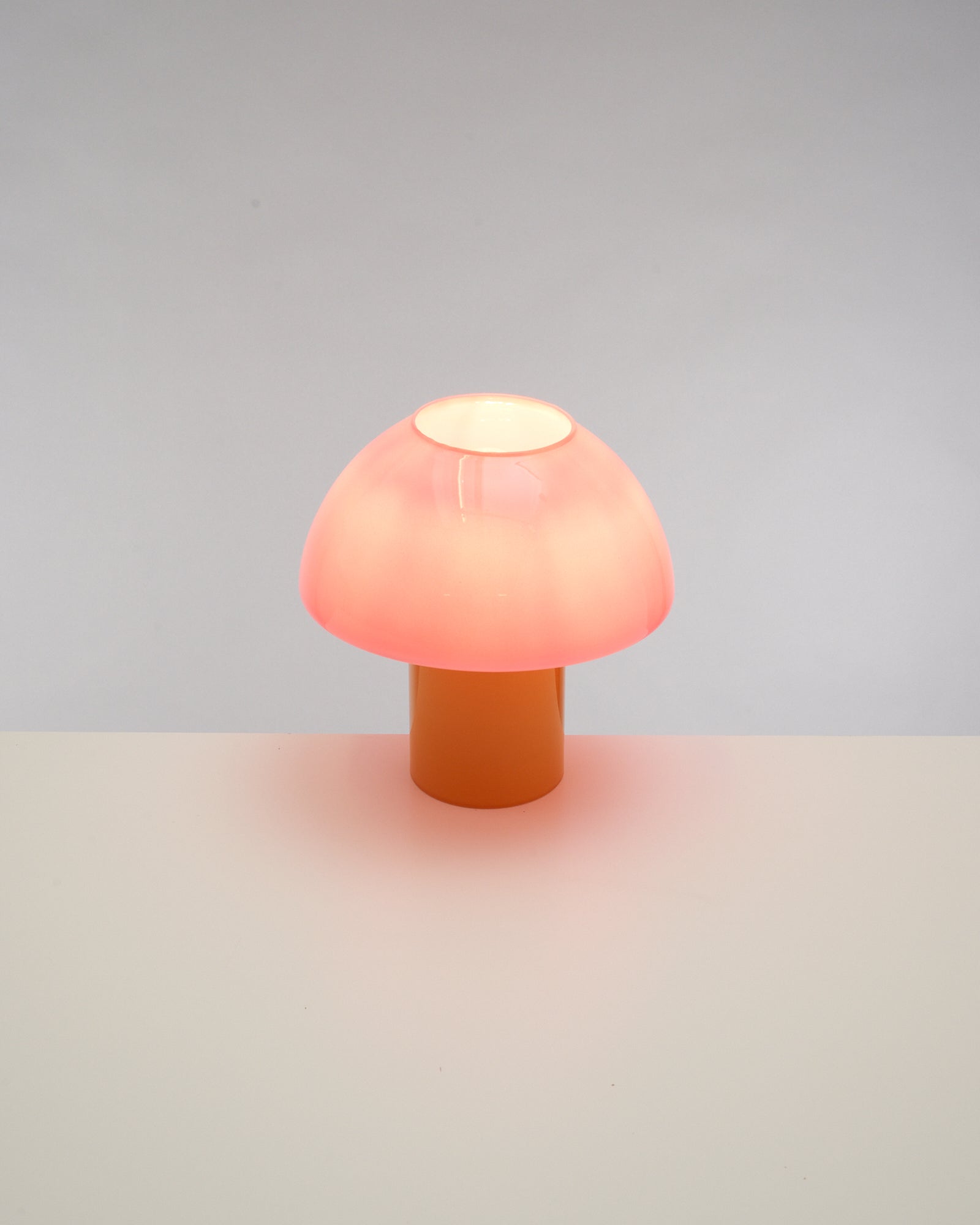 Eine rosa Tischlampe in Pilzform steht auf einer weißen Oberfläche vor grauem Hintergrund.