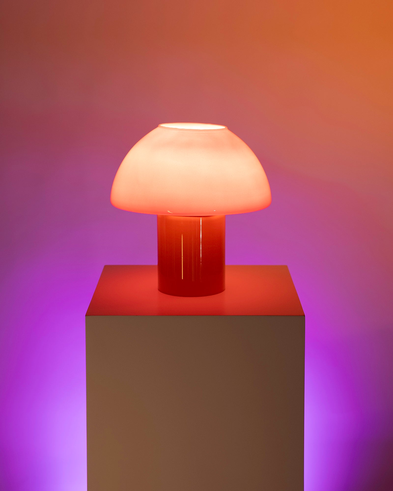 Pilzförmige Lampe in warmem Orange auf einem Sockel vor farbigem Hintergrund.