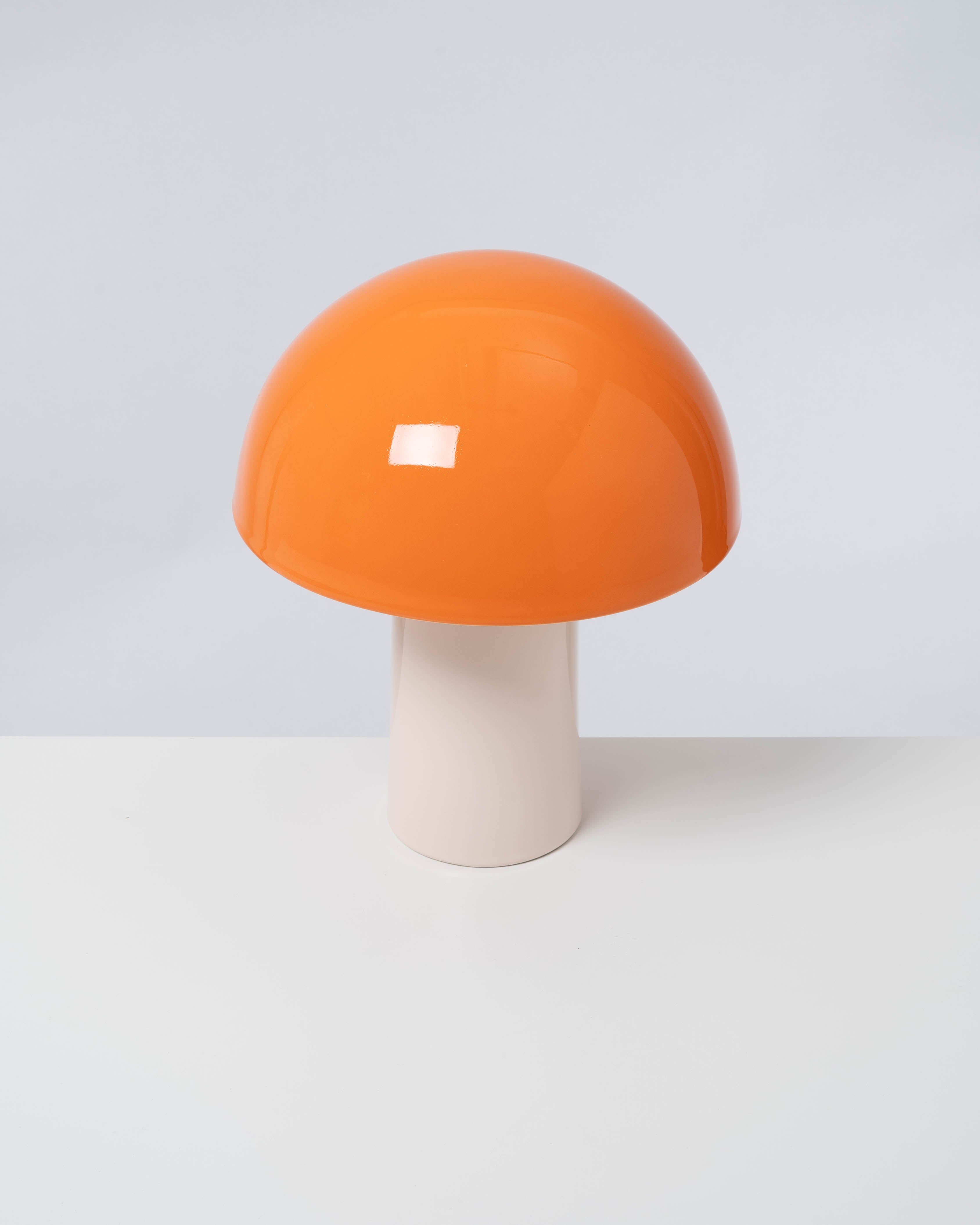 Pilzförmige Lampe mit einem orangefarbenen Schirm und einem weißen Sockel auf einem weißen Untergrund.