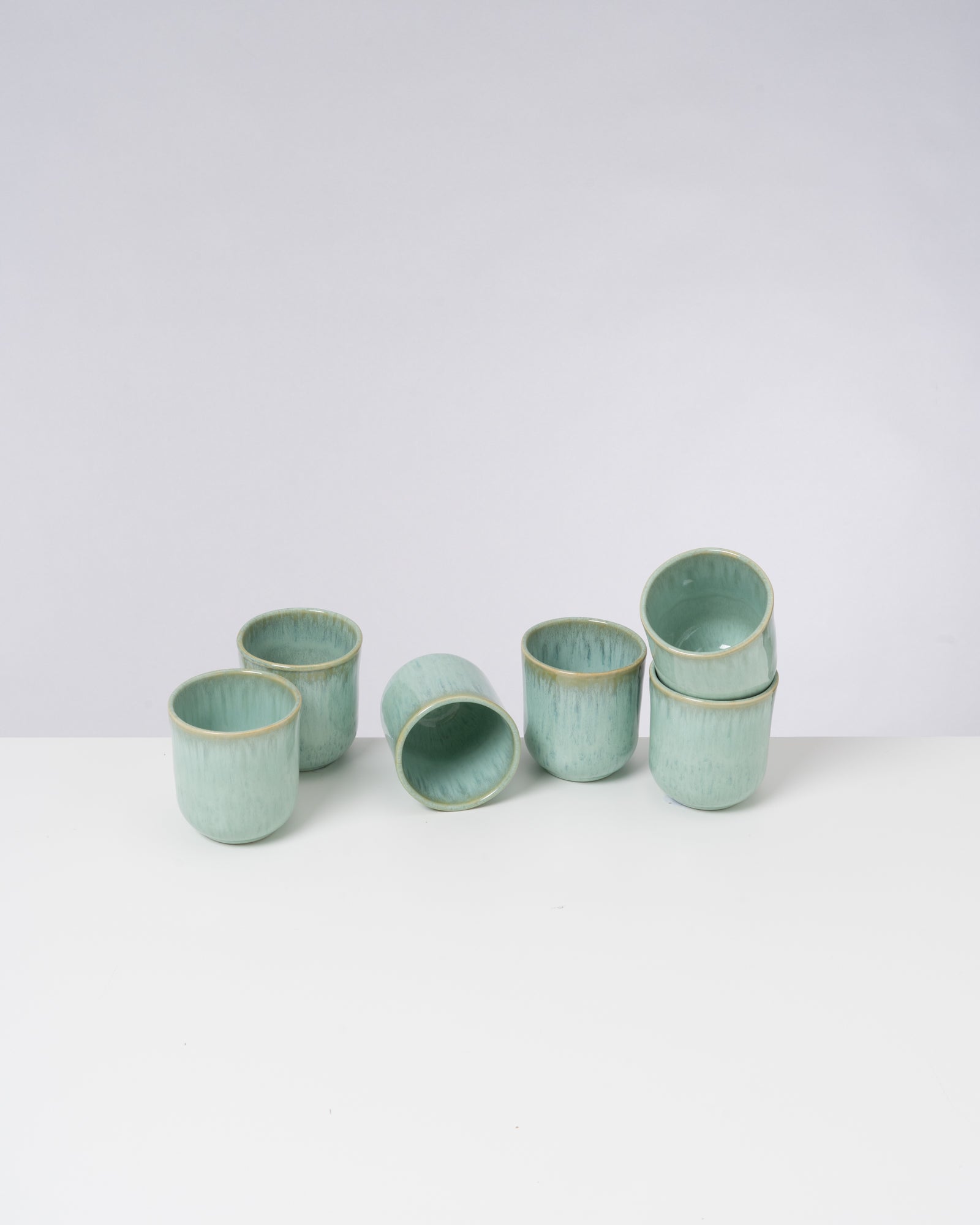 Sieben grüne, keramikähnliche Tassen sind auf einem weißen Tisch arrangiert, einige stehend und eine liegt seitlich.