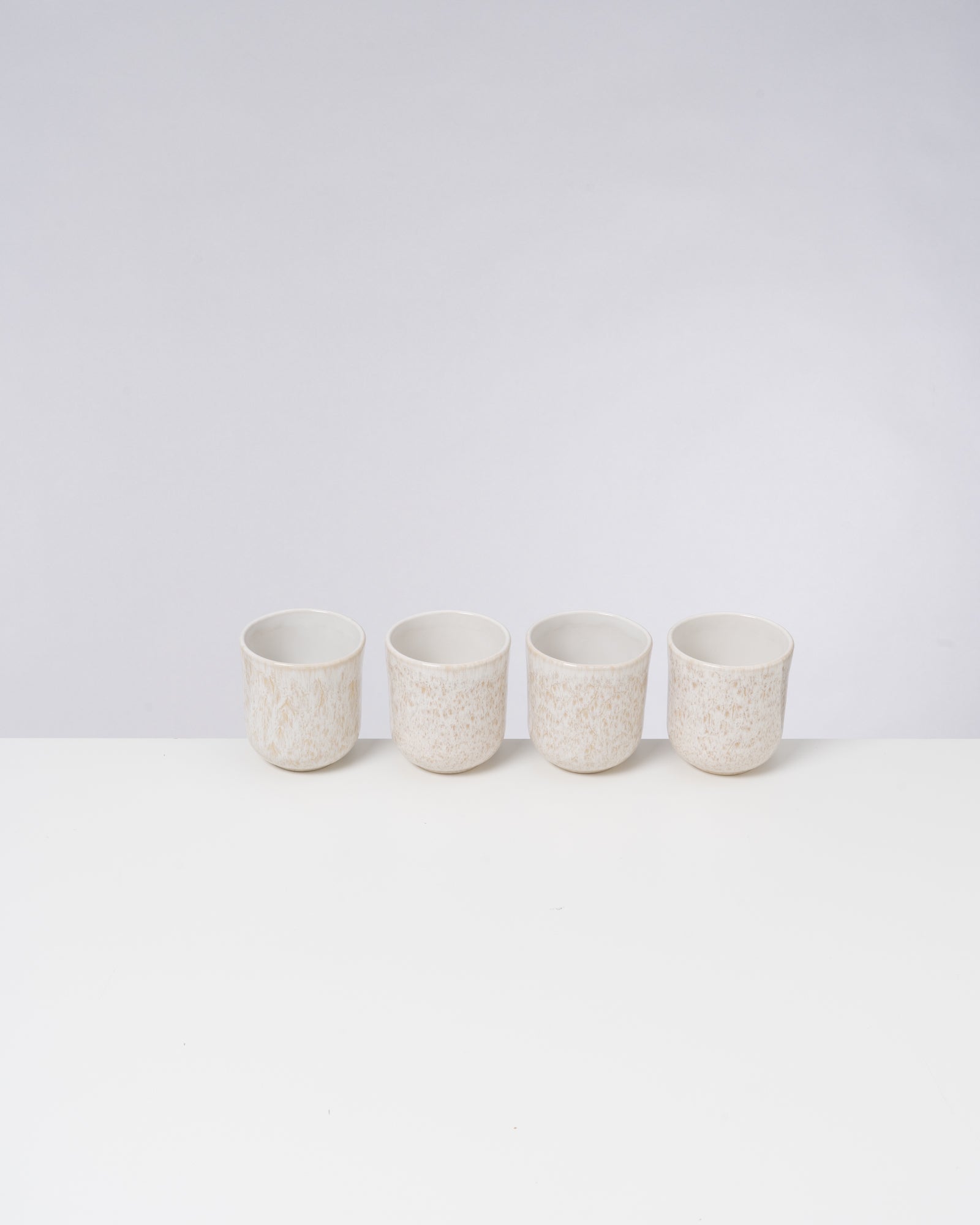 Vier cremefarbene Becher stehen in einer Reihe auf einer weißen Oberfläche vor einem hellgrauen Hintergrund.