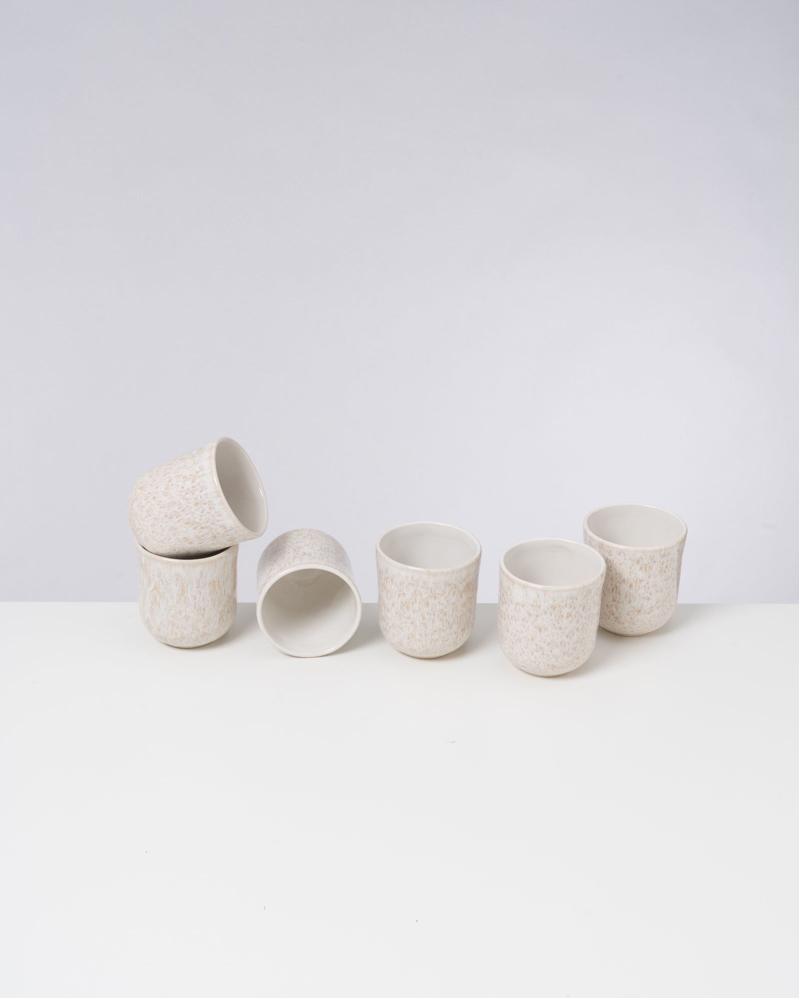 Sechs weiße Keramikbecher mit dezenter Musterung stehen und liegen auf einem weißen Untergrund.