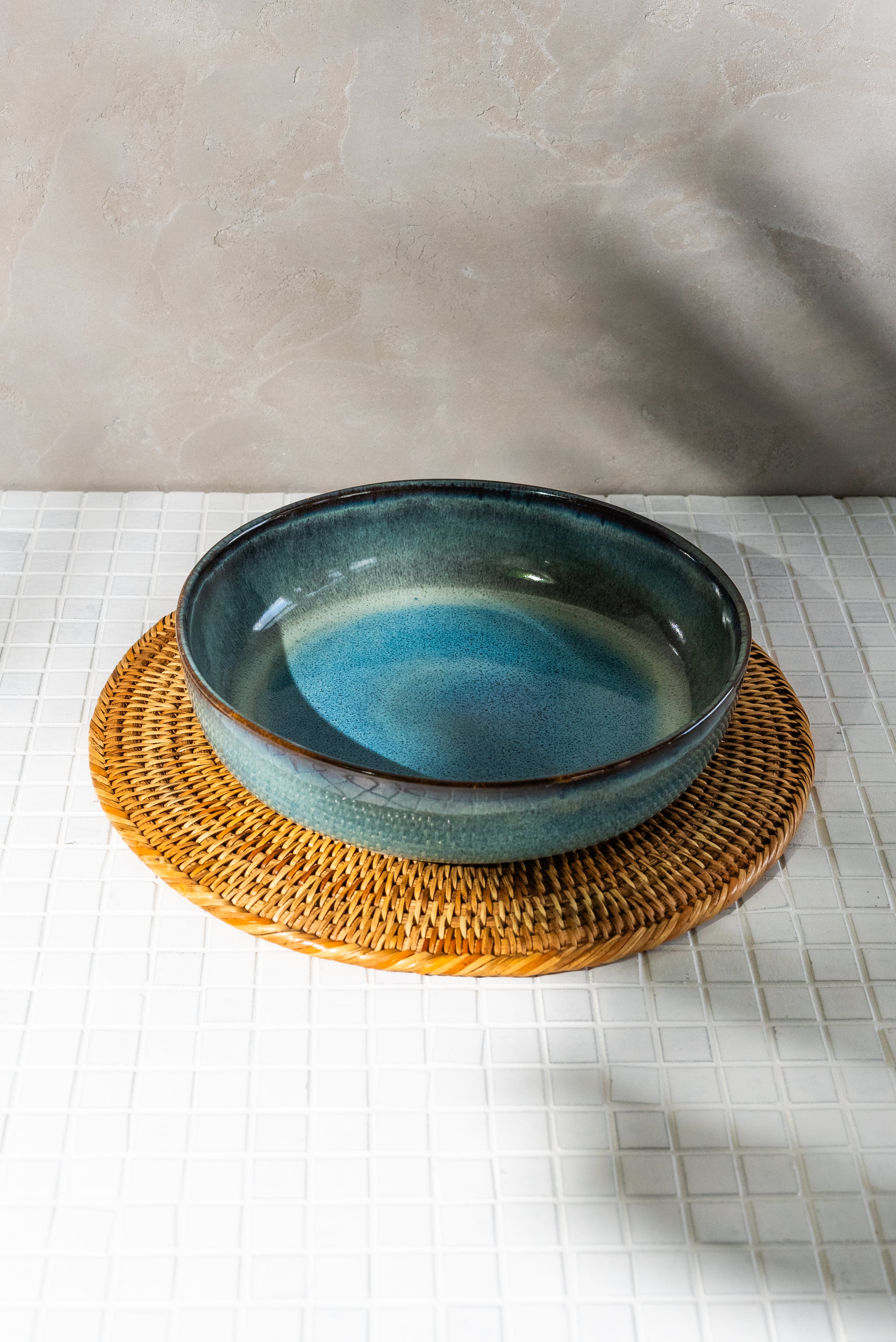 Eine blaue, glasierte Schale steht auf einem runden, geflochtenen Untersetzer auf einem weißen Fliesentisch.