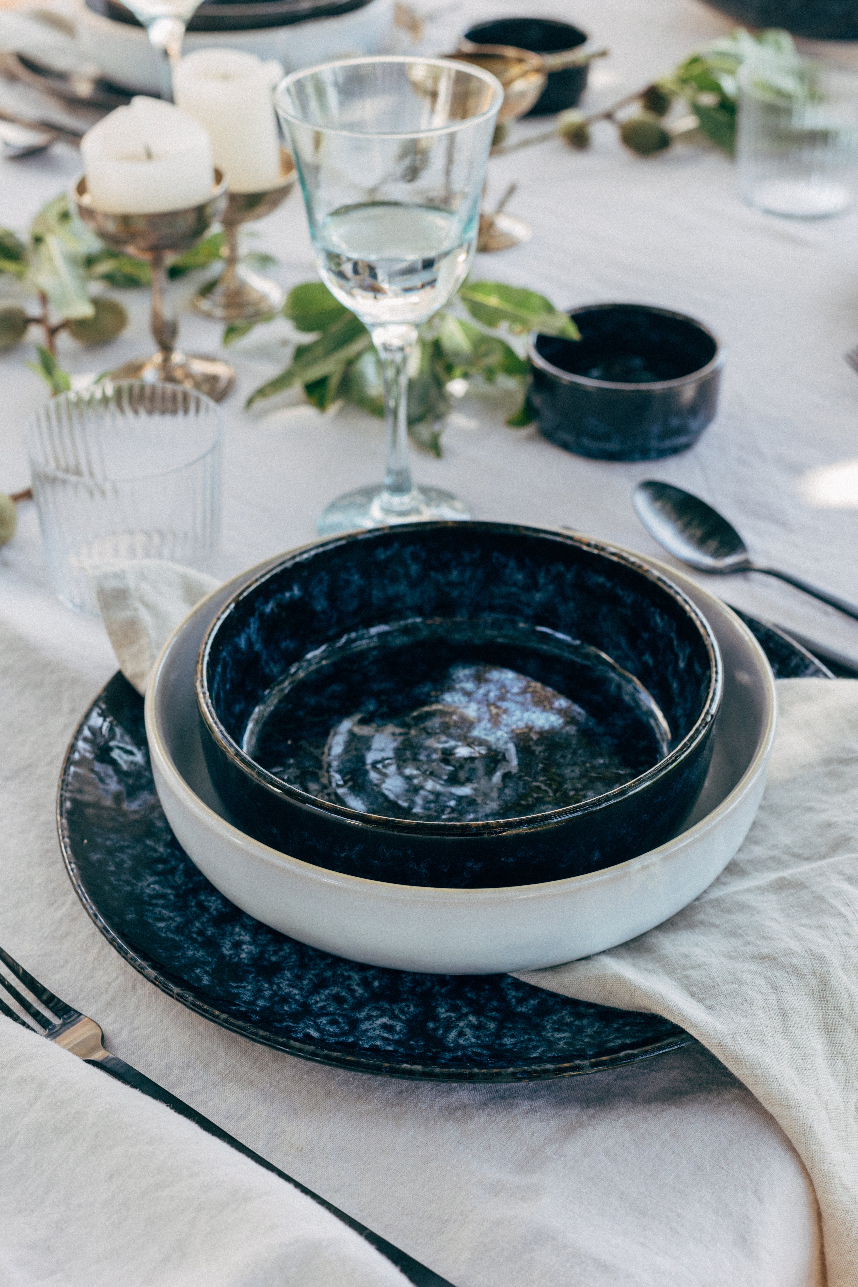 Gedeckter Tisch mit elegantem Geschirr, Gläsern und Kerzen, dekoriert mit grünen Zweigen und Stoffservietten.